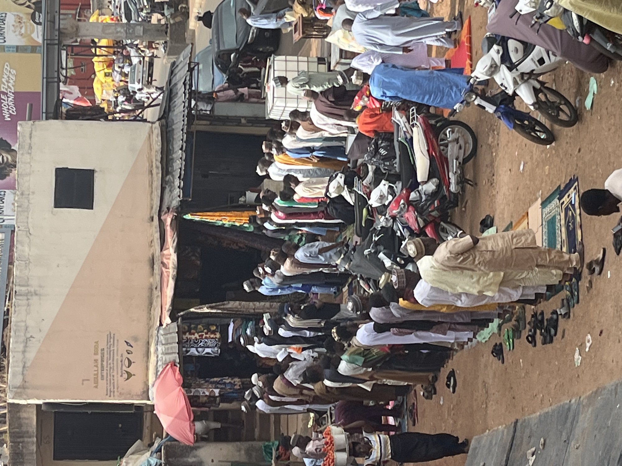 This image depicts an outdoor gathering in Jos, Nigeria, likely a place of worship or a communal event. A group of men are arranged in rows, facing the same direction, suggesting they are engaged in prayer or a similar communal activity. Many of the men are wearing traditional attire, including caps and long robes. In the foreground, several motorcycles are parked, indicating a mode of transportation for some of the attendees. Some individuals are standing near the motorcycles, while others are walking or attending to various tasks. The background shows a bustling street scene with buildings, shops, and market stalls, typical of a Nigerian urban environment. There are also umbrellas and tarpaulins, possibly used for shade or as part of market setups. The overall atmosphere appears to be lively and active, with a sense of community and shared purpose. The presence of motorcycles, market stalls, and the diverse clothing styles all contribute to the authentic depiction of a Nigerian city.
