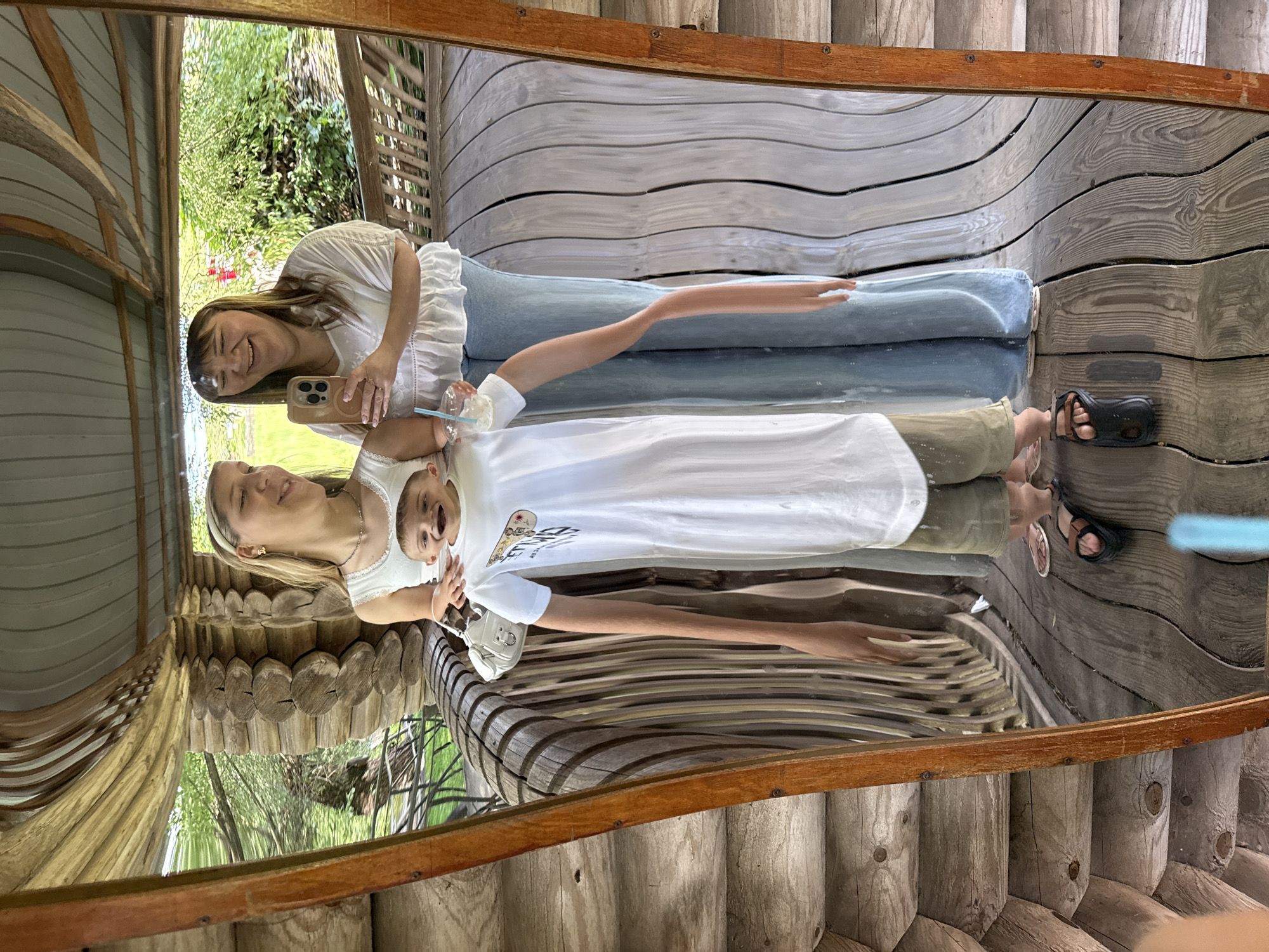 The image shows a distorted reflection in a mirror of two women and a child on a wooden porch. The woman on the left is wearing a white tank top and a headband. The woman on the right is wearing a white embroidered blouse and blue jeans, and is holding a smartphone. Between them, a child in a white t-shirt and olive shorts appears with a distorted, stretched body and face. The reflection reveals a wooden structure with log walls and a wooden floor with warped planks. Green foliage is visible in the background. The location is identified as Kotovo, Russia.