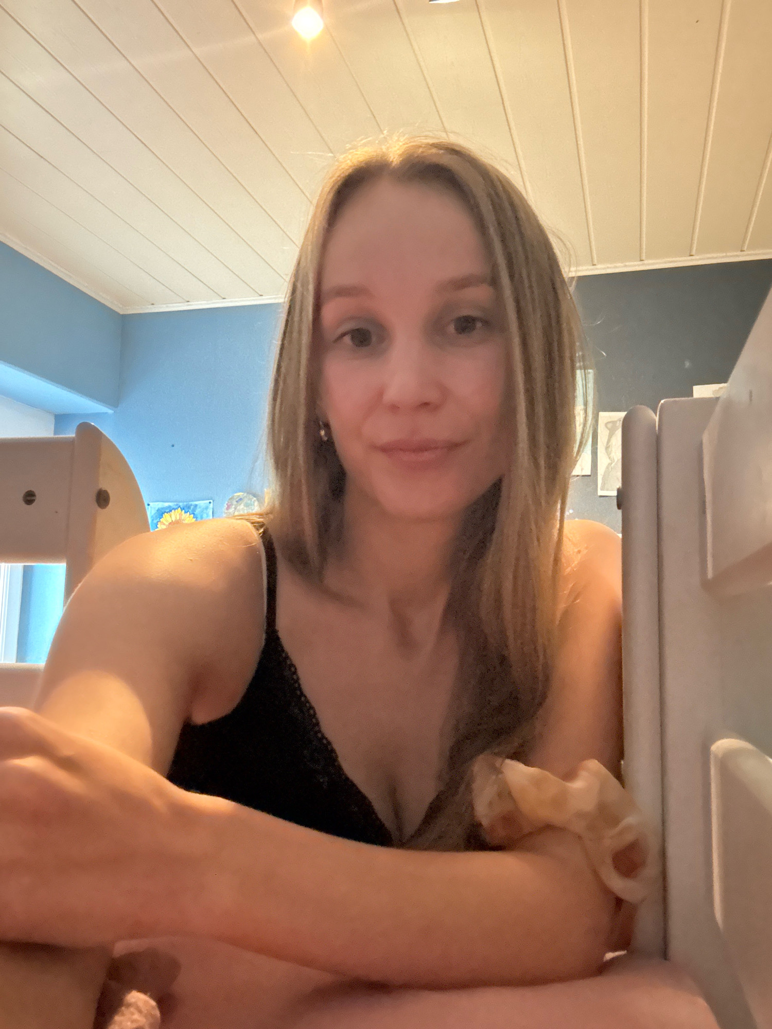 The image is a close-up selfie of a young woman with blonde hair, looking at the camera. She is wearing a black lace-trimmed camisole. She is sitting down, with her arms crossed in front of her. The background is a room with blue walls and a white paneled ceiling. There is a bright light fixture on the ceiling. A light-colored bunk bed or similar furniture is visible to the right of the woman. The overall impression is that of a casual, indoor setting. The image does not provide any specific information that would allow for its location to be identified in the city of Überherrn, Germany.