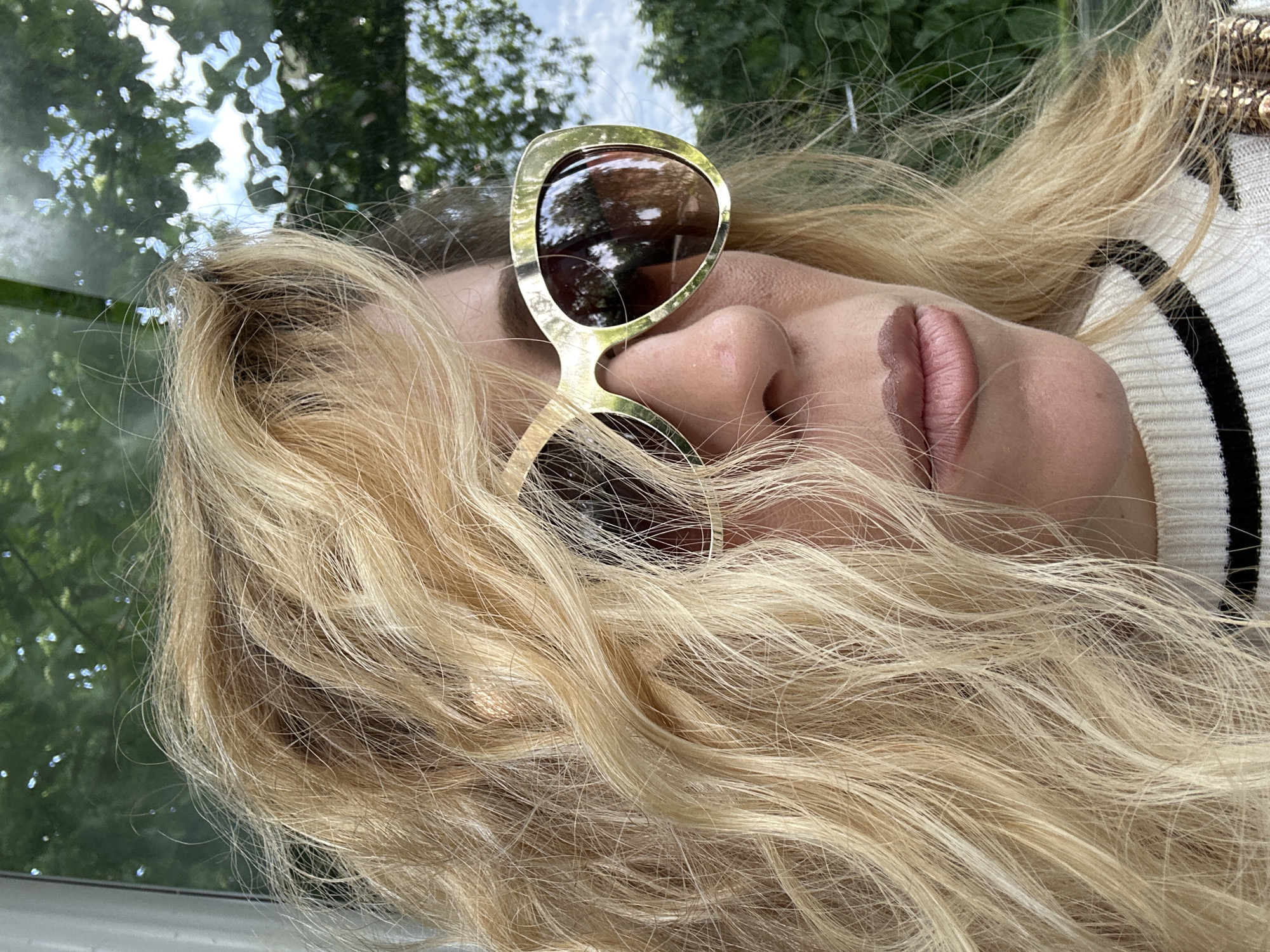 A woman with blonde hair is lying on a surface, likely indoors, with a window showing trees and sky. She is wearing sunglasses with gold frames. Part of a black and white striped shirt or sweater is visible. The location is indeterminable in the city of Dronten, Netherlands.