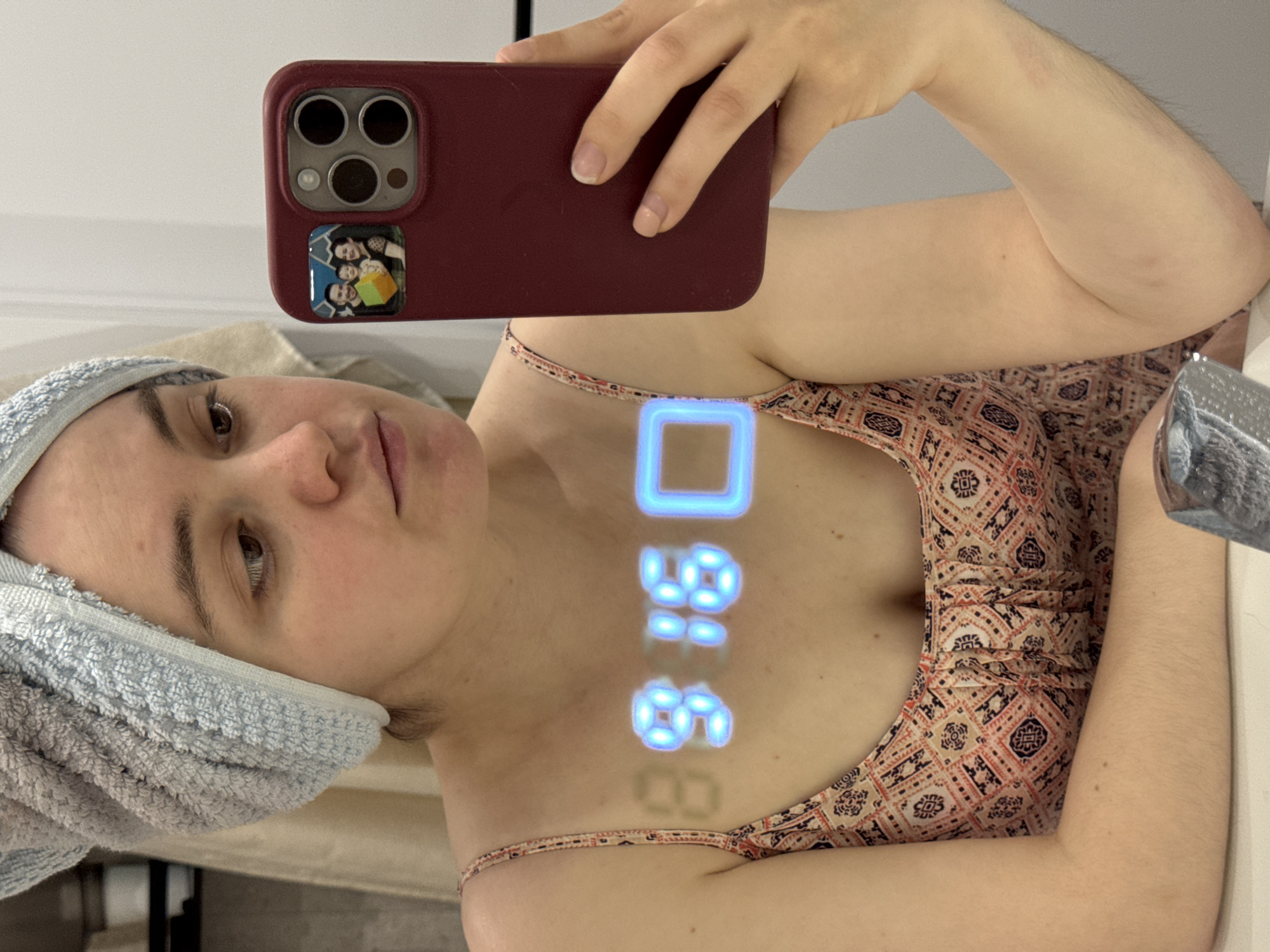 A woman with a towel wrapped around her head takes a selfie in a bathroom mirror, holding a maroon-cased iPhone with a family photo on the back. The bathroom features a patterned top worn by the woman and a chrome faucet. The time displayed on the mirror is 09:58. There is no evidence to suggest a location within Izhevsk, Russia.