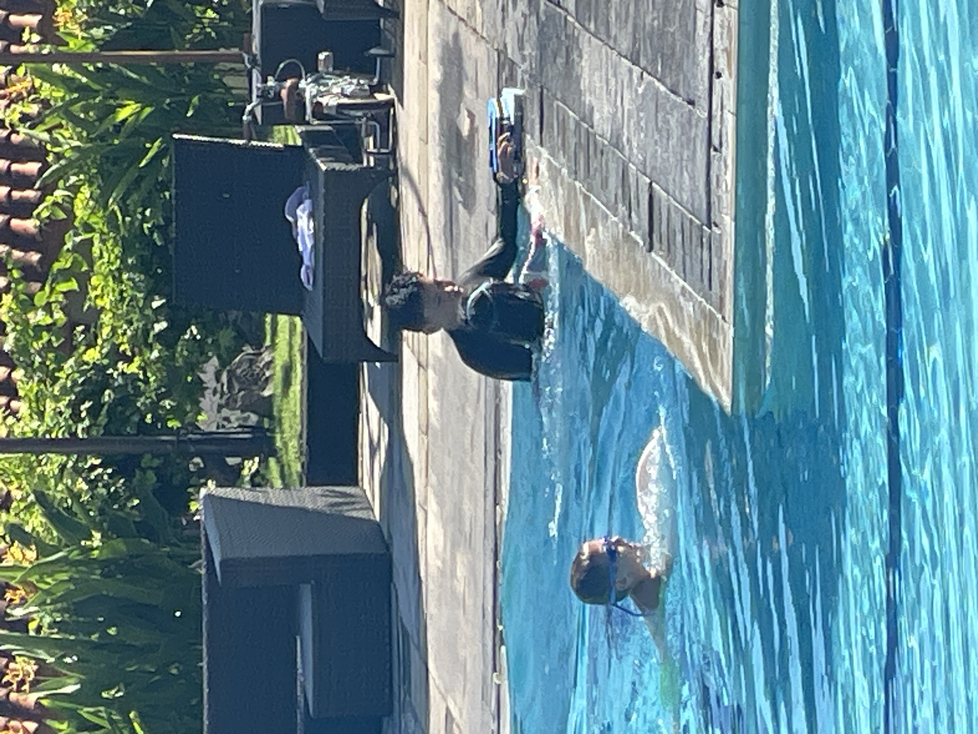 Two children are in a swimming pool in Kuta Selatan, Indonesia. One child, wearing goggles and a green swimming garment, is swimming towards the left side of the frame. The second child, wearing a black wetsuit, is partially submerged near the pool edge, holding a kickboard. The pool is made of bright blue water, with a wooden deck surrounding it. In the background, lounge chairs and lush green foliage are visible.