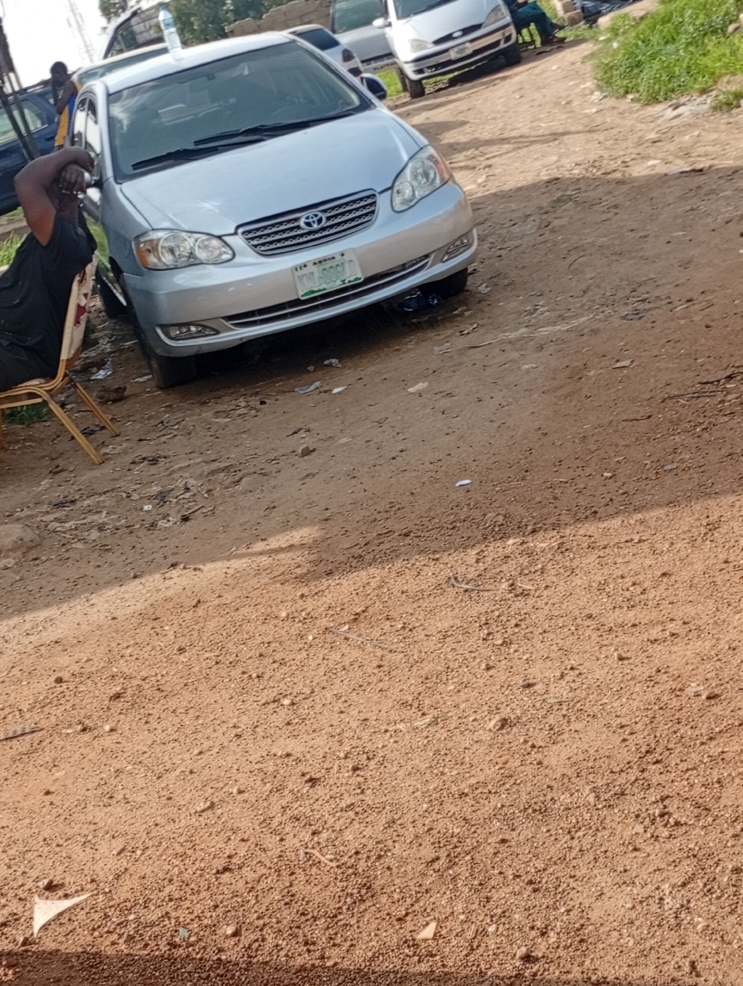 A silver Toyota Corolla is parked on a dirt road in Jos, Nigeria. A person in a black shirt and pants is seated on a gold chair to the left of the car, with their arm across their face. Another person is visible in the background, standing near a dark blue car. The environment is dusty, with some dry grass and scattered debris on the ground.
