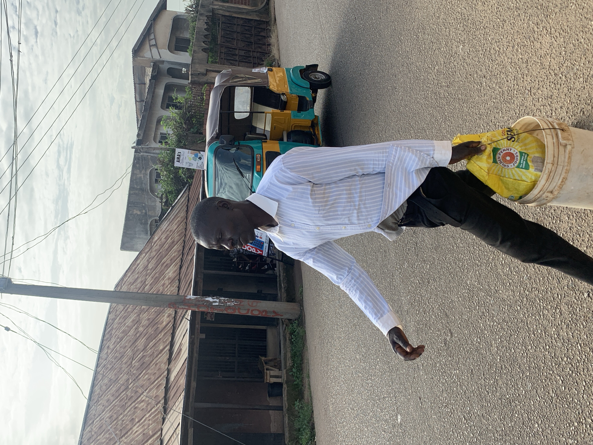 A Black man in a light blue and white striped dress shirt and dark pants walks on a paved road in Jos, Nigeria. He carries a yellow bag with red and green labeling, marked "GOLDEN PENNY FOODS" and "SEM," in a white bucket. Behind him, a teal and yellow three-wheeled auto-rickshaw, or Keke Napep, is parked on the side of the road. The background features buildings with corrugated metal roofs, a utility pole with wires, and some greenery. The sky is overcast.