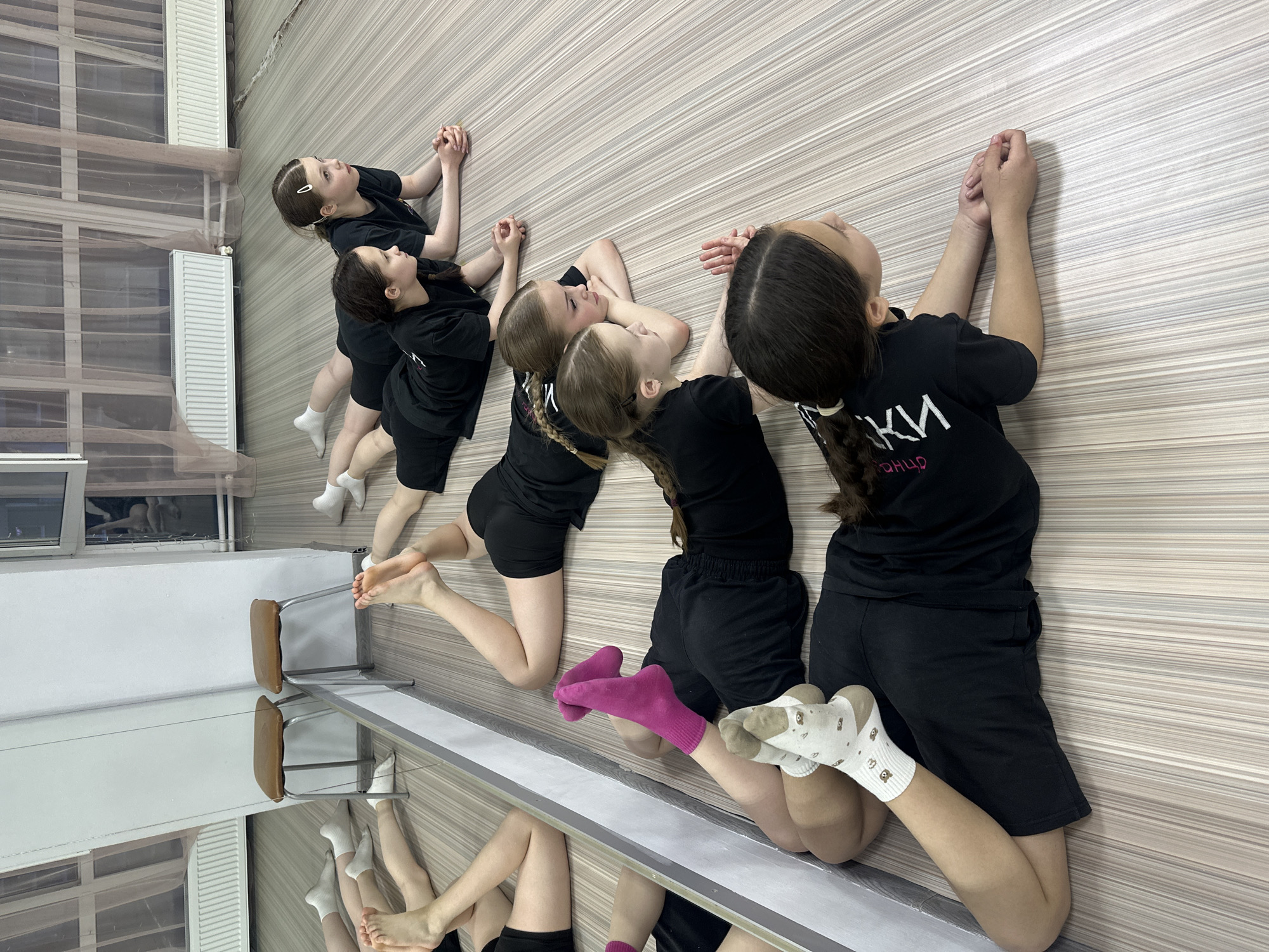 Five girls are lying on their stomachs in a row along a wall, appearing to stretch or relax. They all wear black shirts and black shorts. The girl closest to the viewer has her hair in a ponytail with a white hair tie and is wearing socks with a bear-like pattern. The girl next to her wears a single bright pink sock on one foot, while the other is bare. The next girl has bare feet. The other two girls further down the line are wearing white socks. The location appears to be a dance studio in Votkinsk, Russia, based on lettering on the t-shirt of one of the girls. A mirror reflects the lower portion of the girls' bodies, chairs, and a window. The wall has horizontal stripes.