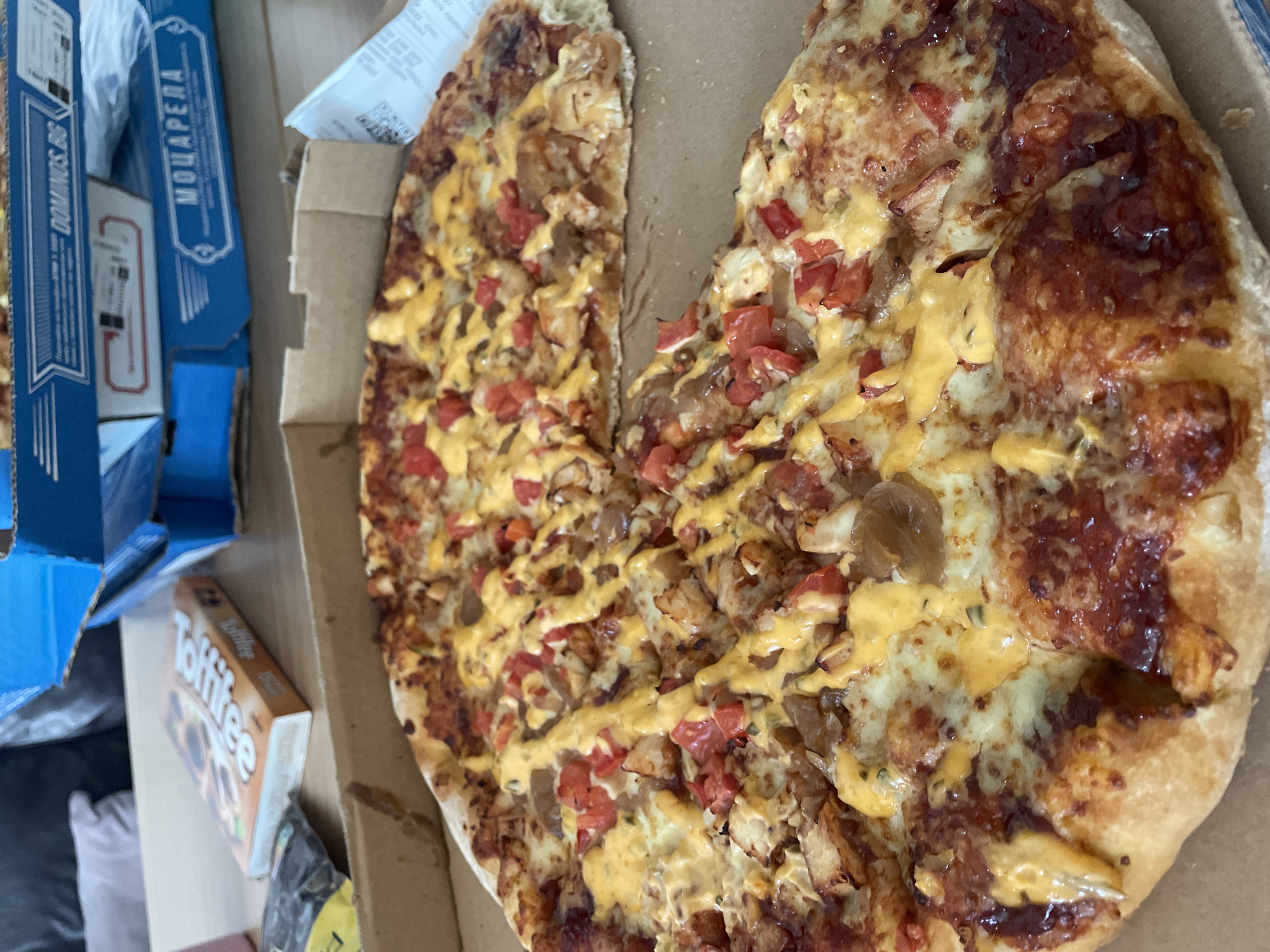 The image shows a partially eaten pizza in a cardboard box, with a Domino's Pizza box and a box of Toffifee candies nearby. The pizza has toppings including cheese, tomatoes, onions, and a yellow sauce. The Domino's Pizza box has "Mozzarella" written on it. The scene is likely indoors. The location of Sofia, Bulgaria, cannot be determined from the given image alone.