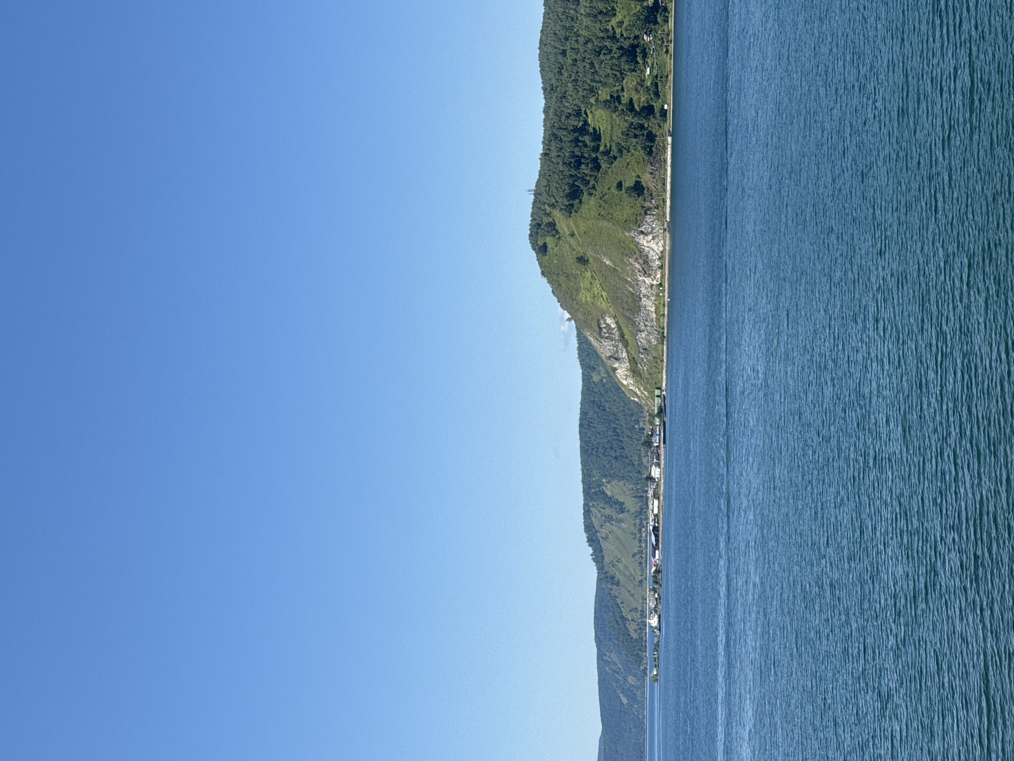 The image depicts a wide, blue body of water, likely a lake or river, with a clear, bright blue sky above. Along the far shore, a line of hills covered in green trees and vegetation stretches across the horizon. A small settlement, appearing as a cluster of buildings with a white-domed structure, is visible nestled at the base of the hills. The scene suggests a natural landscape with a human presence.