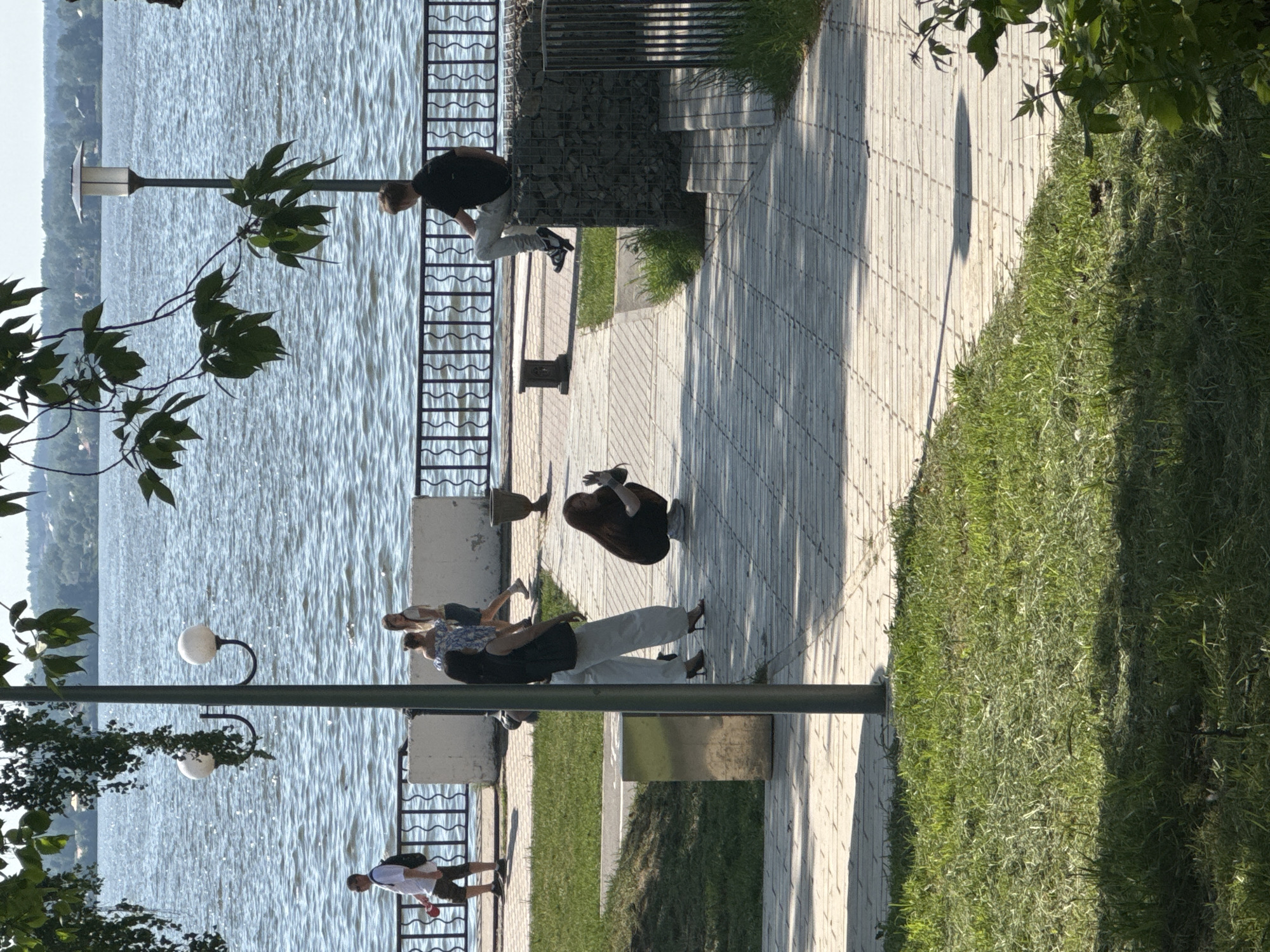 The image shows a lakeside promenade in Votkinsk, Russia, on a sunny day. The promenade is paved with light-colored bricks and lined with a metal fence bordering the water. There are a few people scattered along the promenade: a man in a white shirt and dark shorts is walking, a couple is sitting on a bench, a person with long dark hair is kneeling, and a person in a black shirt is bending over near the fence. The water is light blue and extends to the horizon. A grassy area with trees borders the promenade on the other side. A lamppost with two white globes stands in the foreground, obscuring the scene.
