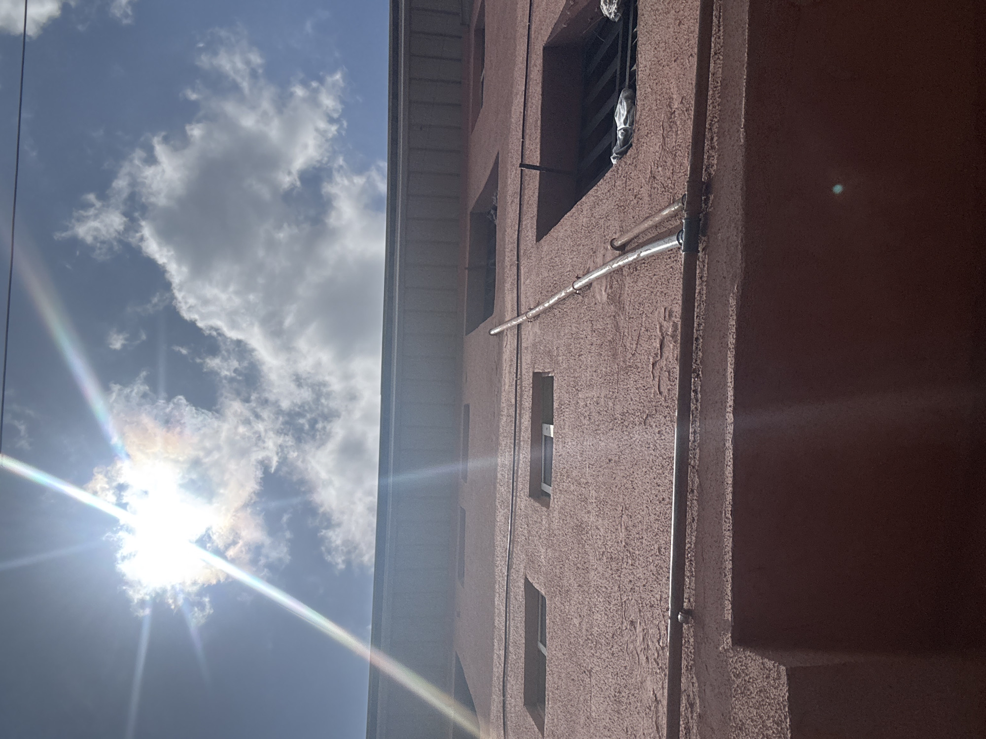 The image shows an exterior view of a multi-story building in Ihiagwa, Nigeria, under a bright, partly cloudy sky. The building is rendered in a reddish-brown color and has multiple windows. A metallic pipe runs vertically along the exterior wall. The sunlight is intense and diffused through clouds, casting a lens flare effect in the sky.