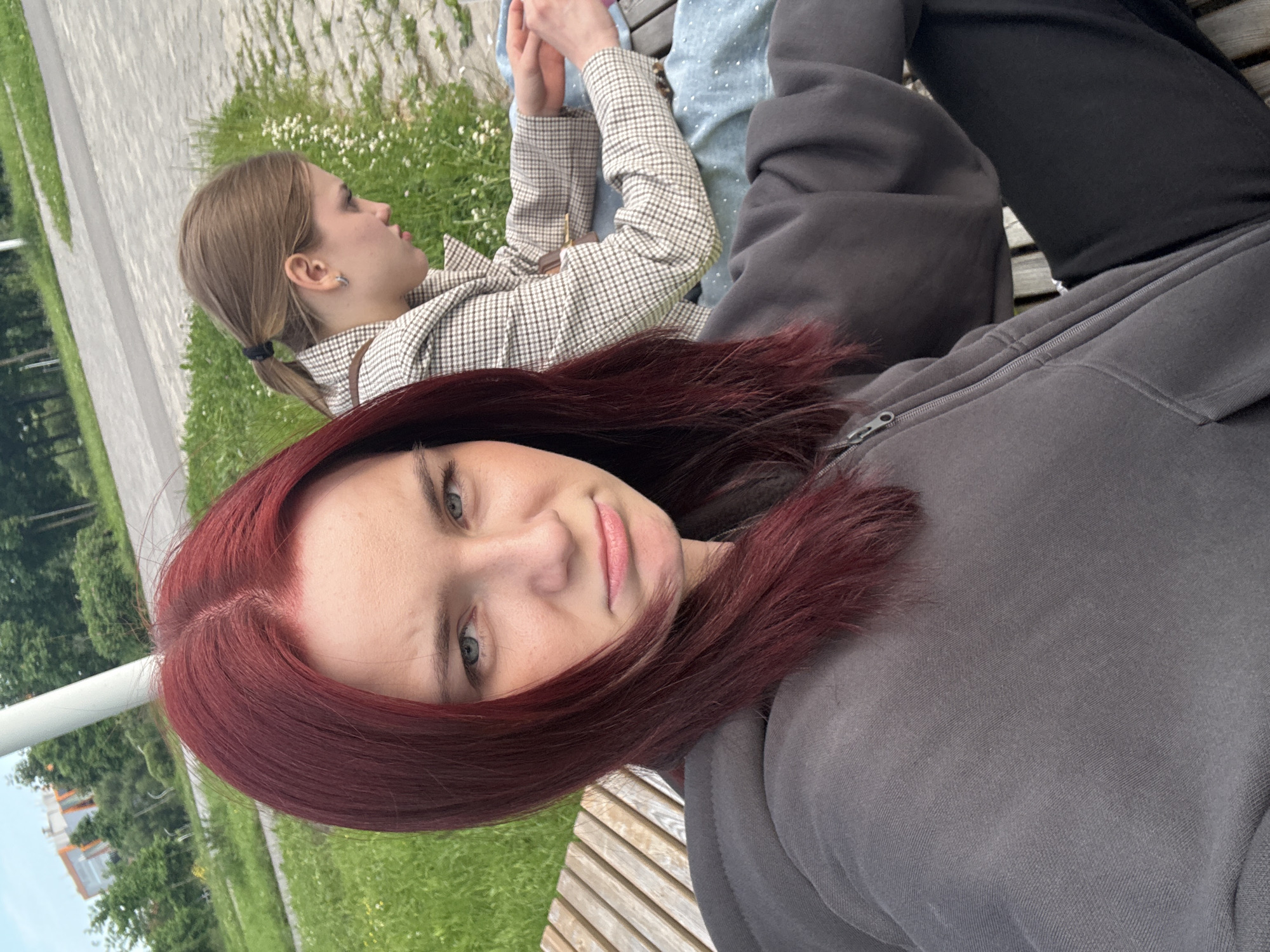 Two young women are sitting on a park bench. The woman in the foreground, facing the camera, has bright red hair and is wearing a grey hooded sweatshirt. She has a neutral to slightly displeased expression on her face. The woman in the background is facing away from the camera, with her head turned to her left, and has her hair pulled back in a ponytail. She is wearing a checkered blazer and a blue top. They are in a park with a paved pathway and green grass in the background, with some buildings and trees visible in the distance. The image appears to be taken outdoors during the day.