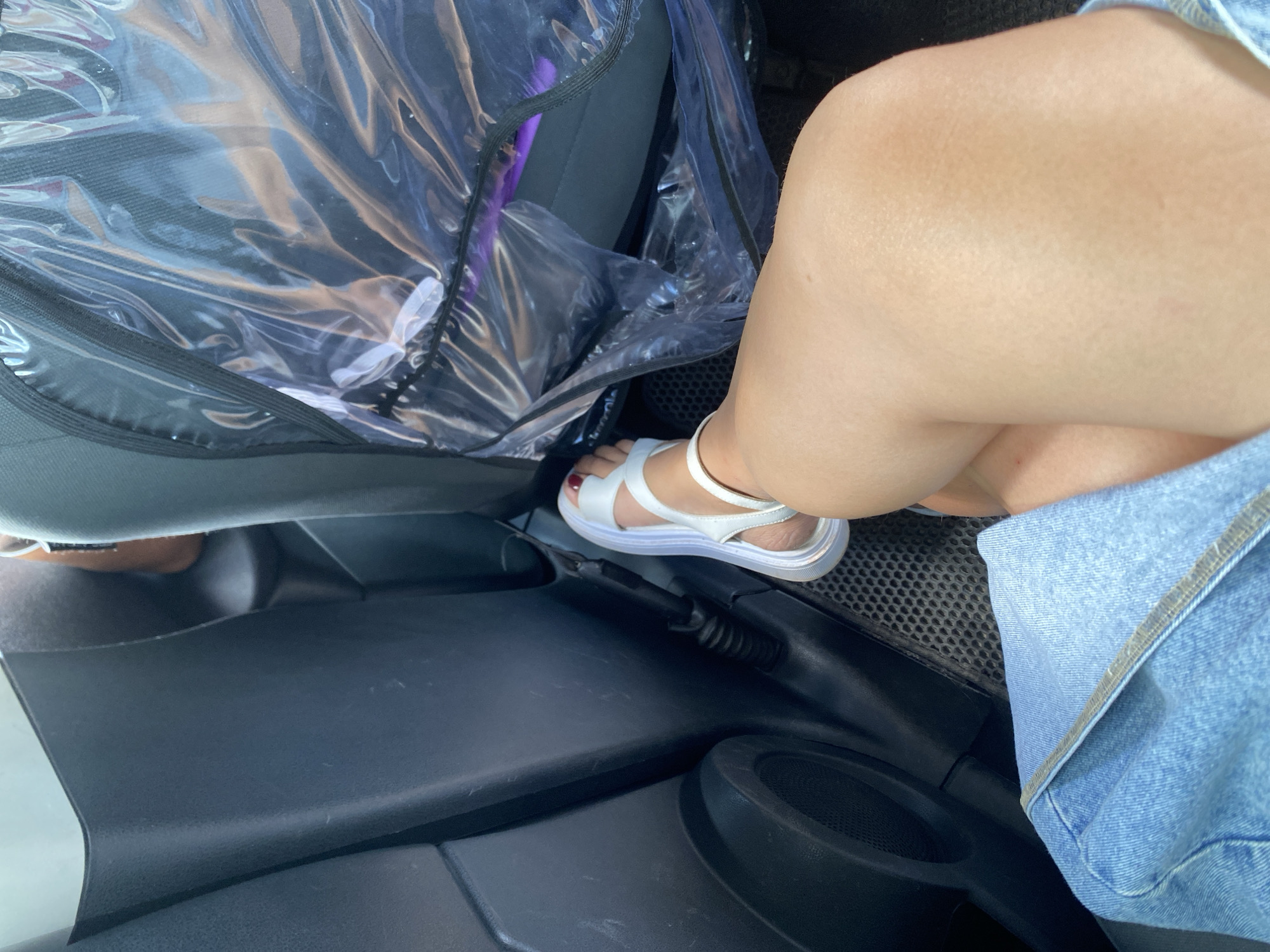 A person's legs are visible inside a vehicle. The person is wearing white sandals and a denim skirt. The interior of the car, including the dark grey plastic paneling and a seat covered in clear plastic, is visible. The floor of the vehicle has a textured mat. The person's right leg is bent at the knee and resting on the left leg, with the foot pointing towards the right. The lighting suggests it is daytime. There is no visible indication of the location being Yuldybayevo, Russia.