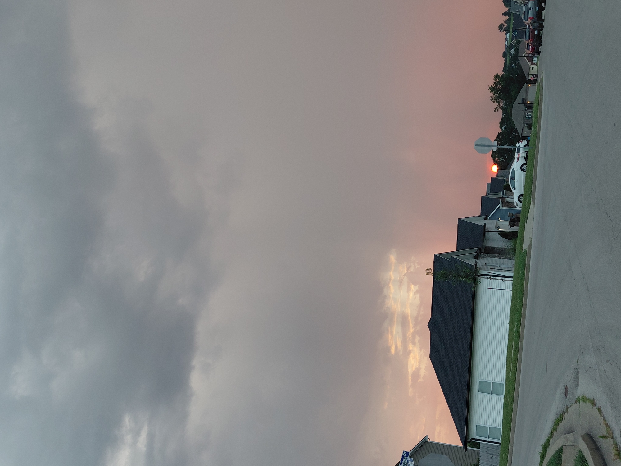 A street corner in Richmond, United States of America, shows a sunset with pink and orange clouds. Several houses are visible, with a white house in the foreground and a white car parked on the street. Some vehicles are parked on the side of the road in the distance. The sky is cloudy, with some darker clouds at the top of the frame.