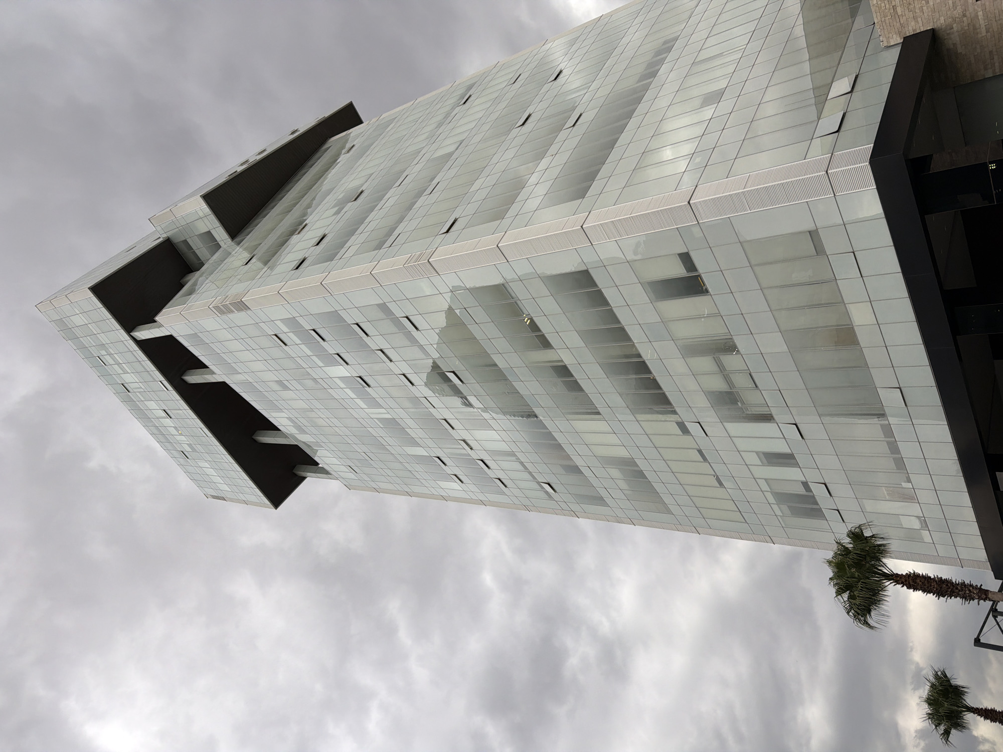 The image presents a low-angle view of a modern, high-rise building in Puebla, Mexico. The building's exterior is predominantly glass, creating a reflective surface that mirrors the cloudy sky above. The building has a boxy design with multiple floors and occasional protruding sections. Palm trees are visible near the base of the building, suggesting an urban environment with tropical vegetation. The sky is overcast, contributing to the overall muted tone of the scene.