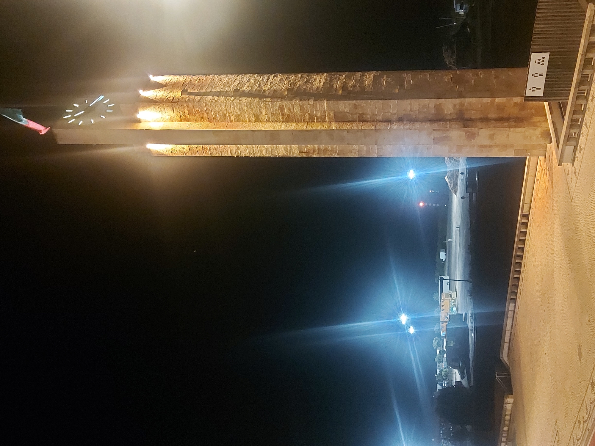 The image depicts the Claridade Clock Tower in Figueira da Foz, Portugal, at night. The tower is illuminated with lights, showcasing its stacked rectangular design. A clock face is visible near the top of the tower, next to a flag. The background features a dark night sky. A street scene is discernible, lit by street lamps. Some vehicles are on the road. The image is taken from a low angle and the scene appears to be captured in vertical orientation.