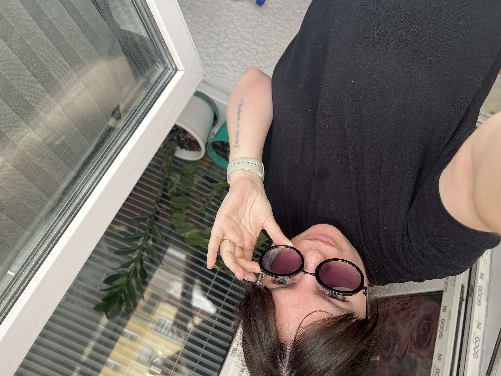 The image depicts an eye-level indoor selfie of a female subject, partially obscured, positioned near a window. The subject has dark brown hair and is wearing a black short-sleeved t-shirt. She is wearing black-framed, round sunglasses with purple-tinted lenses, which she is holding down slightly with her left hand, revealing her blue eyes above the frames. A thin gold band ring is visible on her left ring finger. Her left forearm displays a vertical tattoo with the text "Believe in yourself," and a light-colored silicone watch is worn on the same wrist. The background features a white PVC window frame on the right. Through the window, reflections of an exterior building with light-colored facade and horizontal window elements are visible, along with interior horizontal blinds. A green potted plant, identified as a Zamioculcas zamiifolia (ZZ plant), rests on the windowsill or immediately behind the window. The surface below the window appears to be a light-colored, possibly textured or tiled, sill or floor. The precise location within the city of Noviy, Russia, cannot be confirmed directly from the visual evidence present in the image.