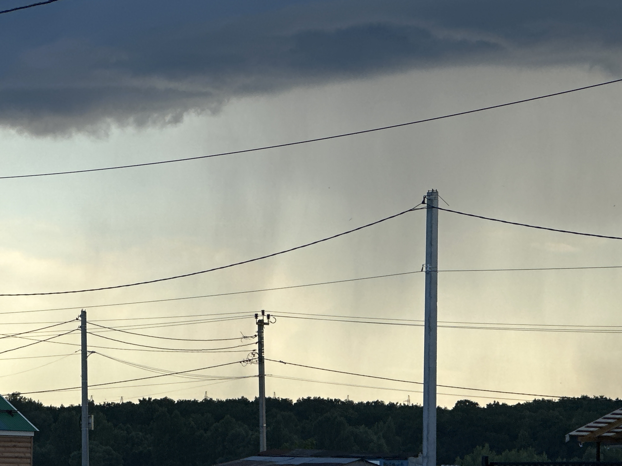 The image shows a view of the sky over a forested area with power lines and utility poles in the foreground. Dark, heavy clouds are present in the upper portion of the sky, with lighter, hazy conditions below, suggesting either a distant rain shower or fog. The treeline is dense and dark green, indicating a natural environment. Several power poles with numerous wires crisscross the scene. In the lower left and right corners, parts of structures are visible, possibly houses or sheds. The overall impression is of an overcast day, possibly with approaching or receding rain, in a semi-rural or suburban setting. Without additional context or visible landmarks, it is impossible to pinpoint the exact location within Ufa, Russia.