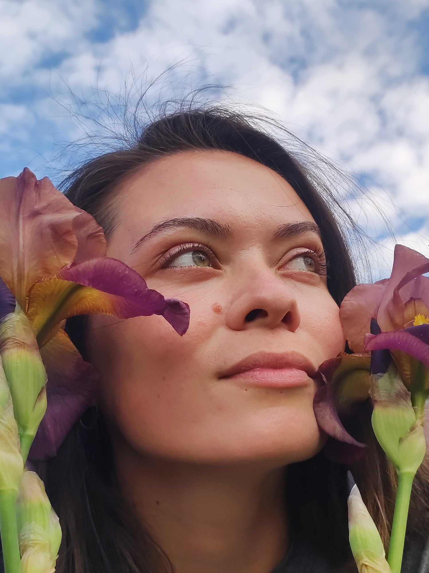 A young woman with long, dark hair is positioned in the foreground of the image, looking upwards and to the right. Her face is framed by two large iris flowers, one on each side. The left iris is partially obscuring her left eye and cheek, while the right iris is positioned near her right eye and cheek. The flowers are a deep purple and brown color with yellow accents. Her skin is fair and appears slightly flushed. Her eyebrows are dark and well-defined. Her eyes are a light green-blue. She has a small mole on her left cheek. The background consists of a blue sky with scattered white clouds. The overall lighting suggests it is daytime. The image appears to be a close-up portrait shot, likely taken outdoors. The provided context of "Фоки, Russia" does not offer any specific visual cues within the image itself to indicate this location.