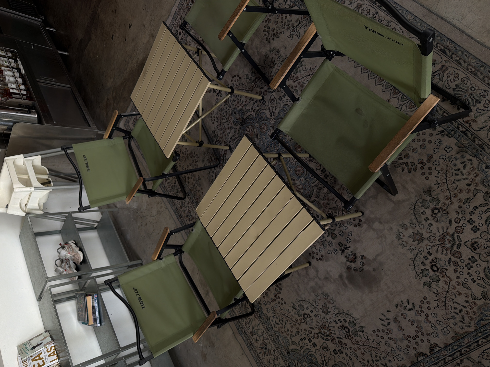 The image shows an indoor scene with two small wooden tables surrounded by four green camping chairs. The tables have a slatted surface and appear to be lightweight. The chairs are also green with wooden armrests. The setup seems to be on a patterned rug, partially visible in the bottom right corner.
In the background to the left is a metal shelving unit containing various items, including a white bin, shoes, books, and cleaning supplies. Further back, a metal counter can be seen.
The location information (North Kuta, Indonesia) is not verifiable from the image.