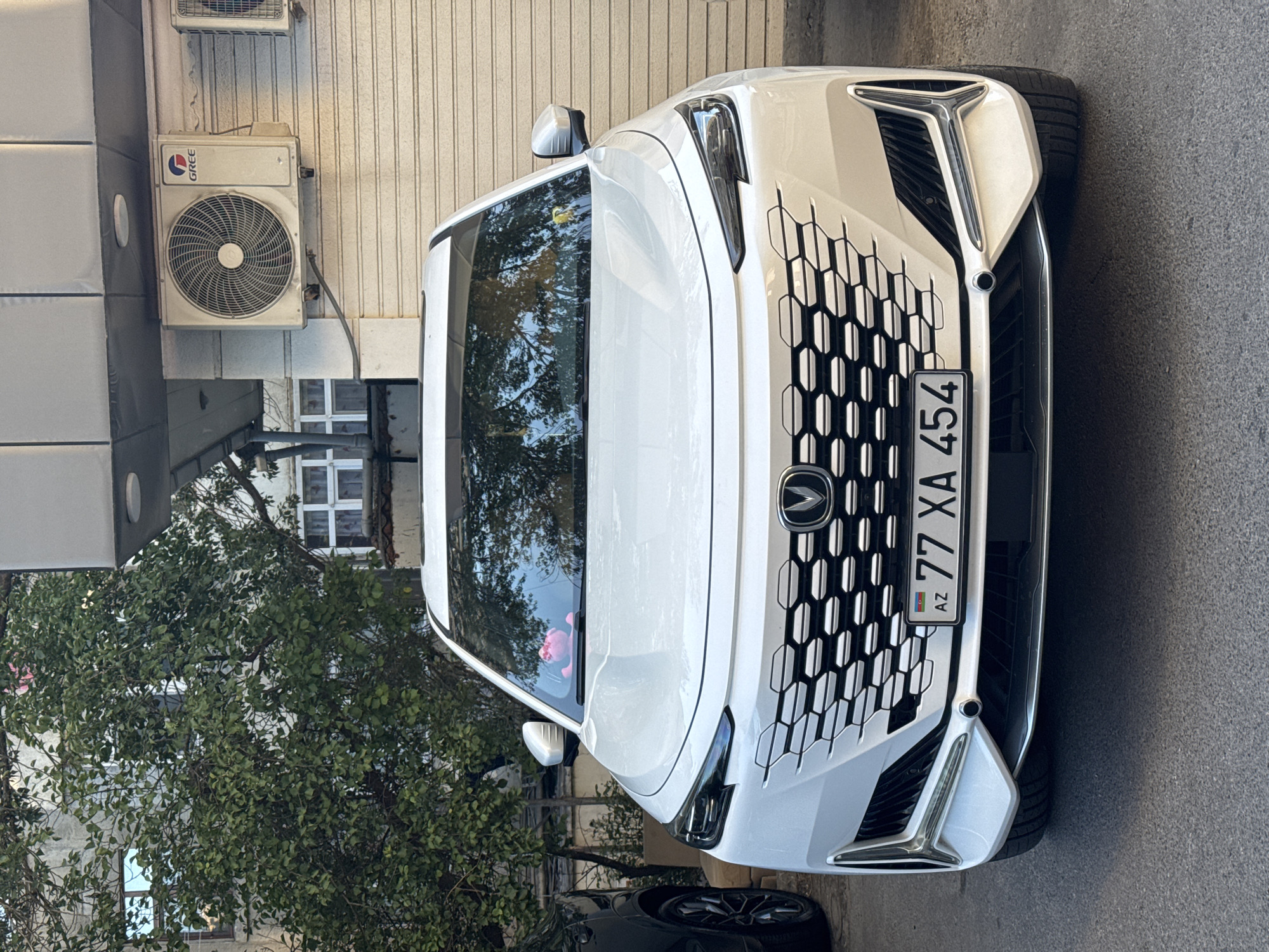 A white sedan is parked on an asphalt street in front of a building with an air conditioning unit mounted on its exterior. The car has a honeycomb-patterned grille with the brand logo in the center and an Azerbaijani license plate that reads "77 XA 454". A pink stuffed animal is visible on the dashboard, reflected in the windshield. The building has windows with white frames and a corrugated metal roll-up door. A green tree with dense foliage is partially obscuring the building. The scene is consistent with an urban setting in Baku, Azerbaijan.