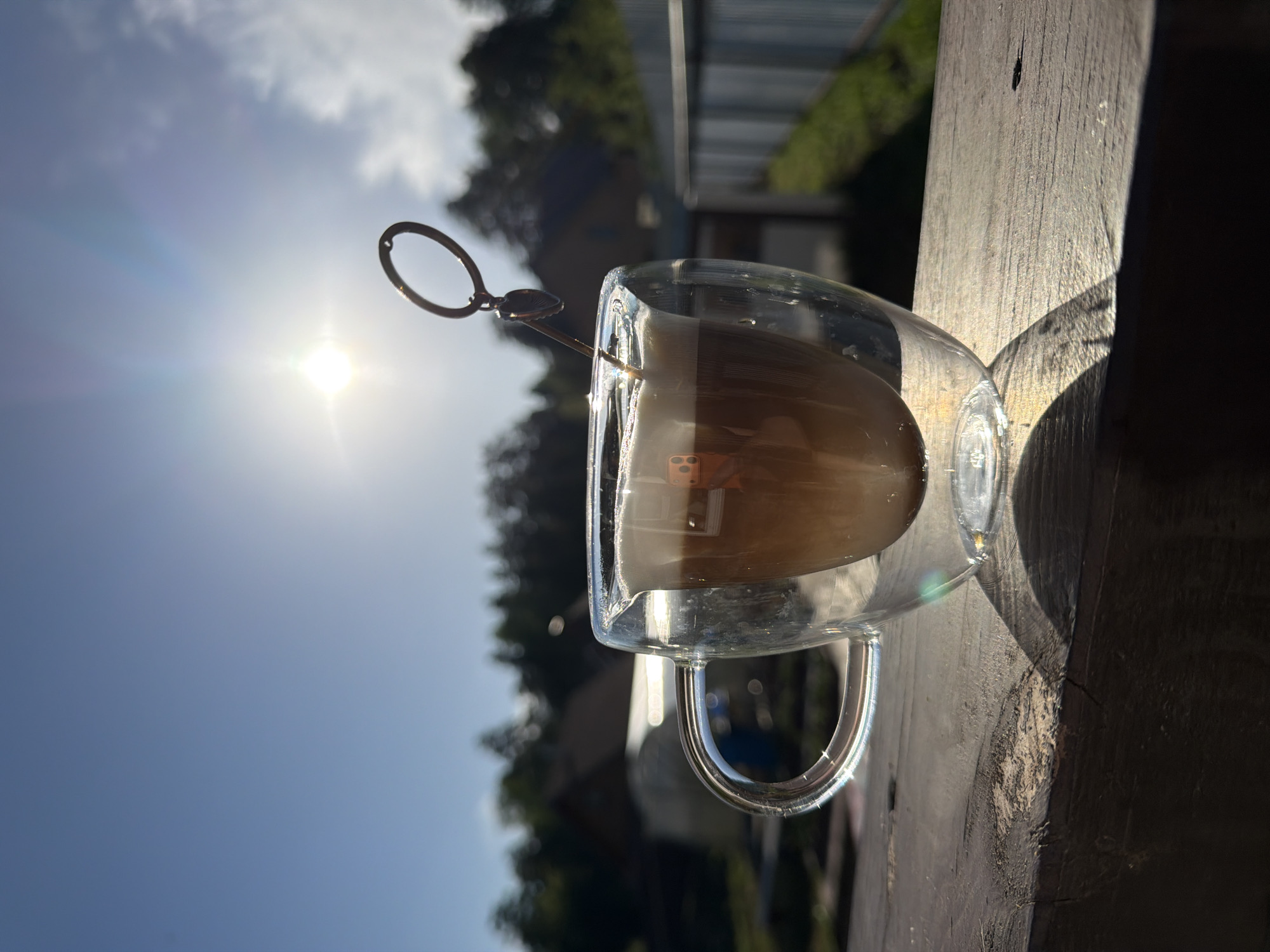 A close-up shot depicts a clear, double-walled glass mug filled with a light brown liquid, possibly coffee or tea. A metal stirrer with a circular loop at the end rests in the mug. The mug sits on a weathered wooden surface, likely a deck or railing, with the sun shining brightly in the background. The background includes foliage, glimpses of buildings, and a fence. The scene seems to be outdoors during daylight. There is no verifiable indication that the location is in the city of Chaikovsky, Russia.