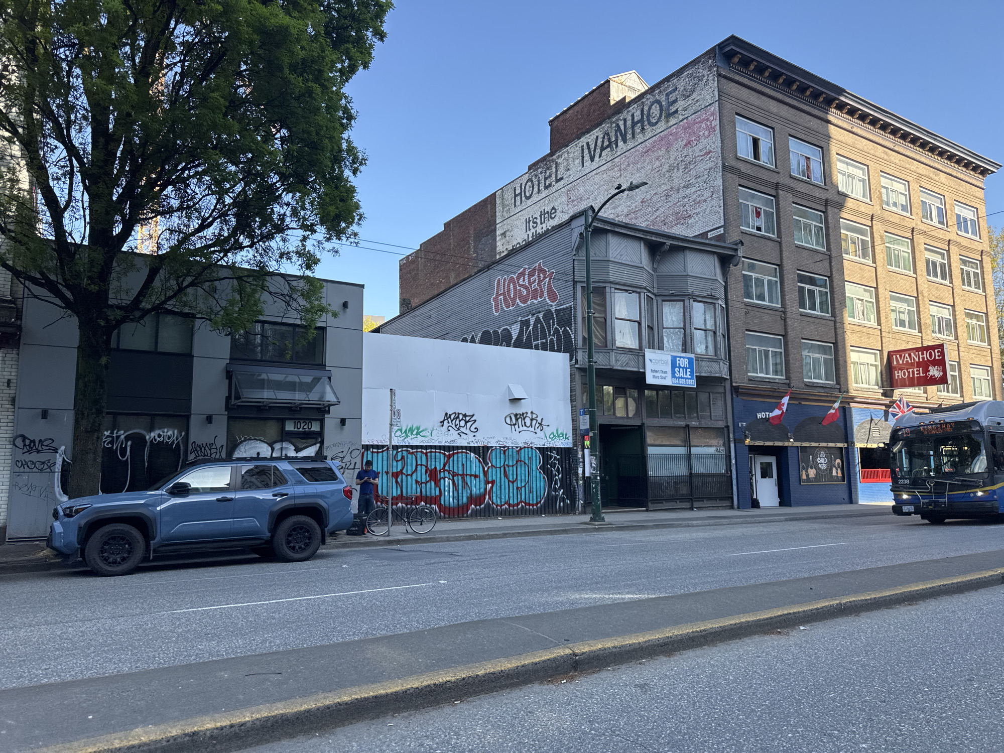 The image depicts a street scene in Vancouver, Canada, showcasing a mix of architectural styles and urban elements. A blue SUV is parked in front of a grey building with graffiti. Next to the SUV, a person is standing with a bicycle, looking at their phone. Adjacent to them is a building boarded up with graffiti. "FOR SALE" sign is displayed on the next building. Further down the street stands the Ivanhoe Hotel, partially obscured by the building next to it. A bus is seen on the right side of the image, approaching the camera. The sky is clear and blue.