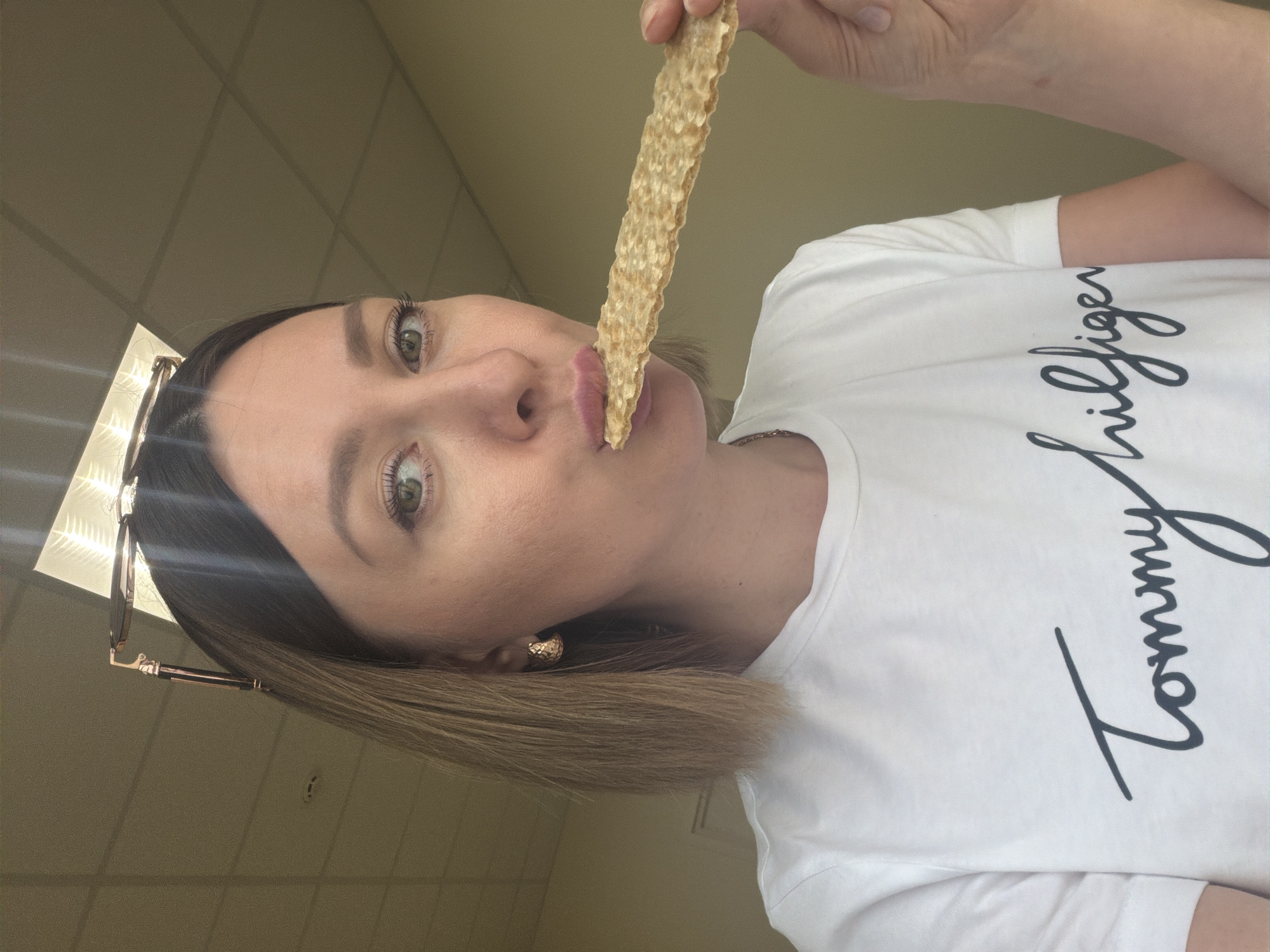 The image presents a close-up, angled shot of a woman indoors, taken from a slightly upward perspective. She has light brown, shoulder-length hair, with sunglasses resting on top of her head. Her green eyes are wide and focused directly at the camera, and she wears subtle makeup. She is dressed in a white t-shirt that prominently features the brand name "Tommy Hilfiger" in black script across the chest.
The woman is holding a long, golden-brown, textured snack, resembling a cracker or crispbread, in her right hand. She is bringing the snack to her mouth, appearing either about to take a bite or having just tasted it. A patterned, gold-toned earring is visible in her left ear.
The background reveals an indoor setting with a plain, light-colored wall and a ceiling composed of grid-patterned acoustic tiles, typical of commercial or institutional environments. Bright indoor lighting is present, with noticeable reflections in her eyes and the lenses of her sunglasses. The overall scene captures a casual, posed moment in Rostov-on-Don, Russia.