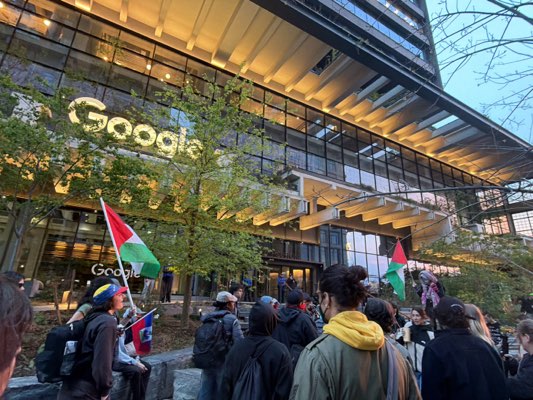 The image shows a group of people gathered in front of a building with "Google" signage. The building appears to be a modern office structure with glass windows and architectural elements. Some people in the group are holding flags, including what seems to be a Palestinian flag. The overall environment suggests a possible protest or demonstration taking place in front of the Google building. The exact location within Hoboken, United States is not discernible from the image.