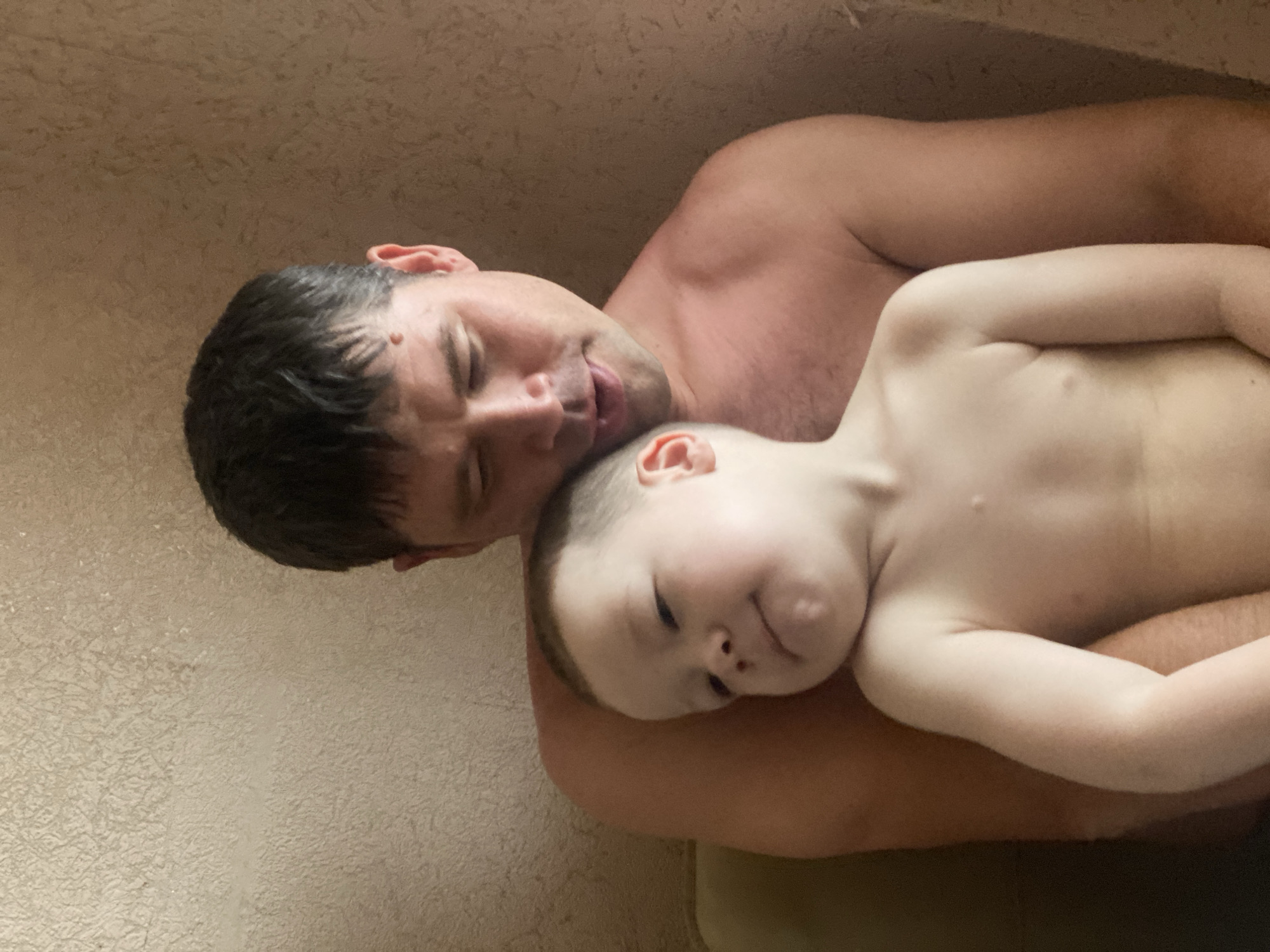 An adult male is depicted embracing a young child in an indoor setting, identified as Sochi, Russia. The male, appearing to be in his 30s or 40s, has short, dark hair and a noticeable mole superior to his right eyebrow. He is bare-chested, revealing tanned skin, and is looking downwards at the child with his mouth slightly ajar. He holds the child securely against his left side. The child, a toddler with fair skin and light hair, is also bare-chested and rests their head against the adult's left shoulder. The child's eyes are partially closed, and their gaze is directed to the left of the frame, with a subtle, slight upturn at the corners of their mouth. The background consists of a textured wall, appearing to be stucco or rough plaster, varying in color from light beige on the left to a warmer tan on the right. A small portion of what appears to be green or light brown furniture is visible behind the adult male's left shoulder.