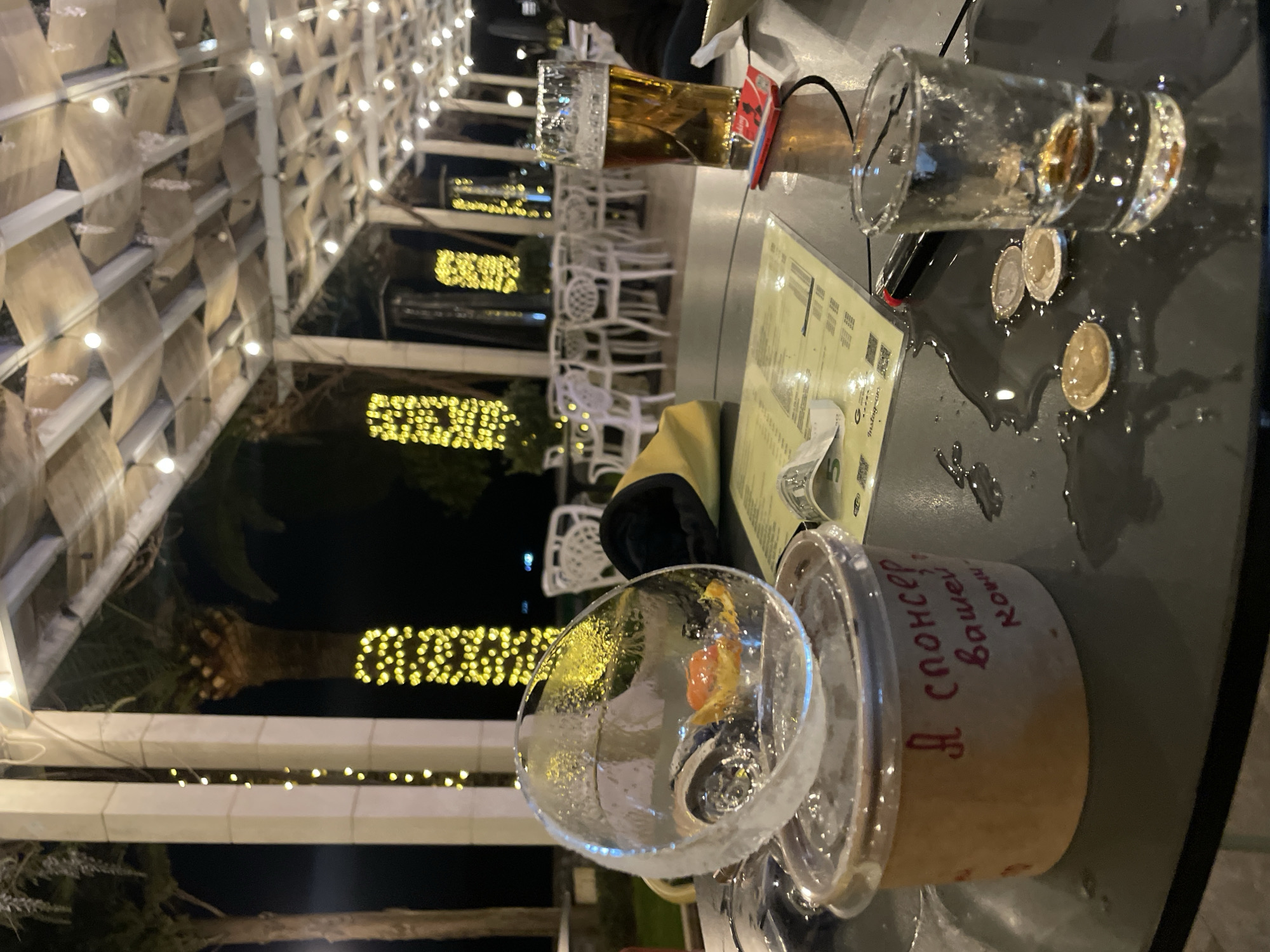 The scene is set at an outdoor bar or restaurant in Rafailovići, Montenegro, illuminated by warm fairy lights and strings of lights spelling out words in Cyrillic script.
The primary focus is on a table with a dark gray surface. Several glasses and containers occupy the space. There's a tall beer glass partially filled with beer, an empty shot glass, and a plastic container filled with ice and a piece of fruit. The container has Cyrillic text written in red ink. Spilled liquid surrounds the shot glass along with a few coins. A menu lies partially visible on the table.
In the background, white chairs and tables suggest a bustling environment. Lights highlight the structure of the outdoor seating area, creating a contrast with the darker background of trees.
