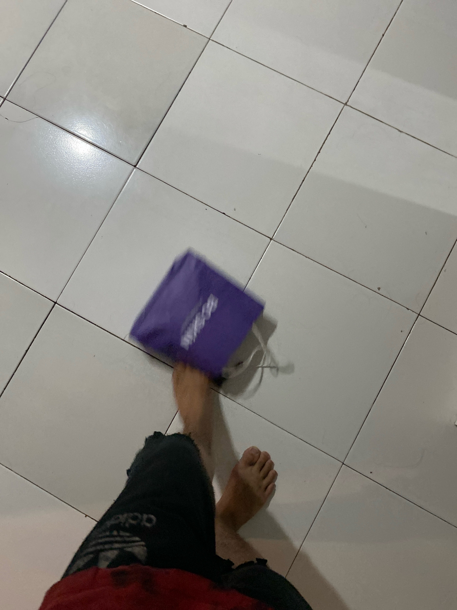 The image shows a person's feet and legs standing on a tiled floor. The person is wearing black pants with an Adidas logo and a red shirt. A small purple bag with white text is being held or dropped by the person, appearing slightly blurred due to motion. The floor is made of light-colored square tiles with dark grout lines. The scene appears to be indoors. There are no specific landmarks or elements that would allow for pinpointing a location within Klurak, Indonesia.