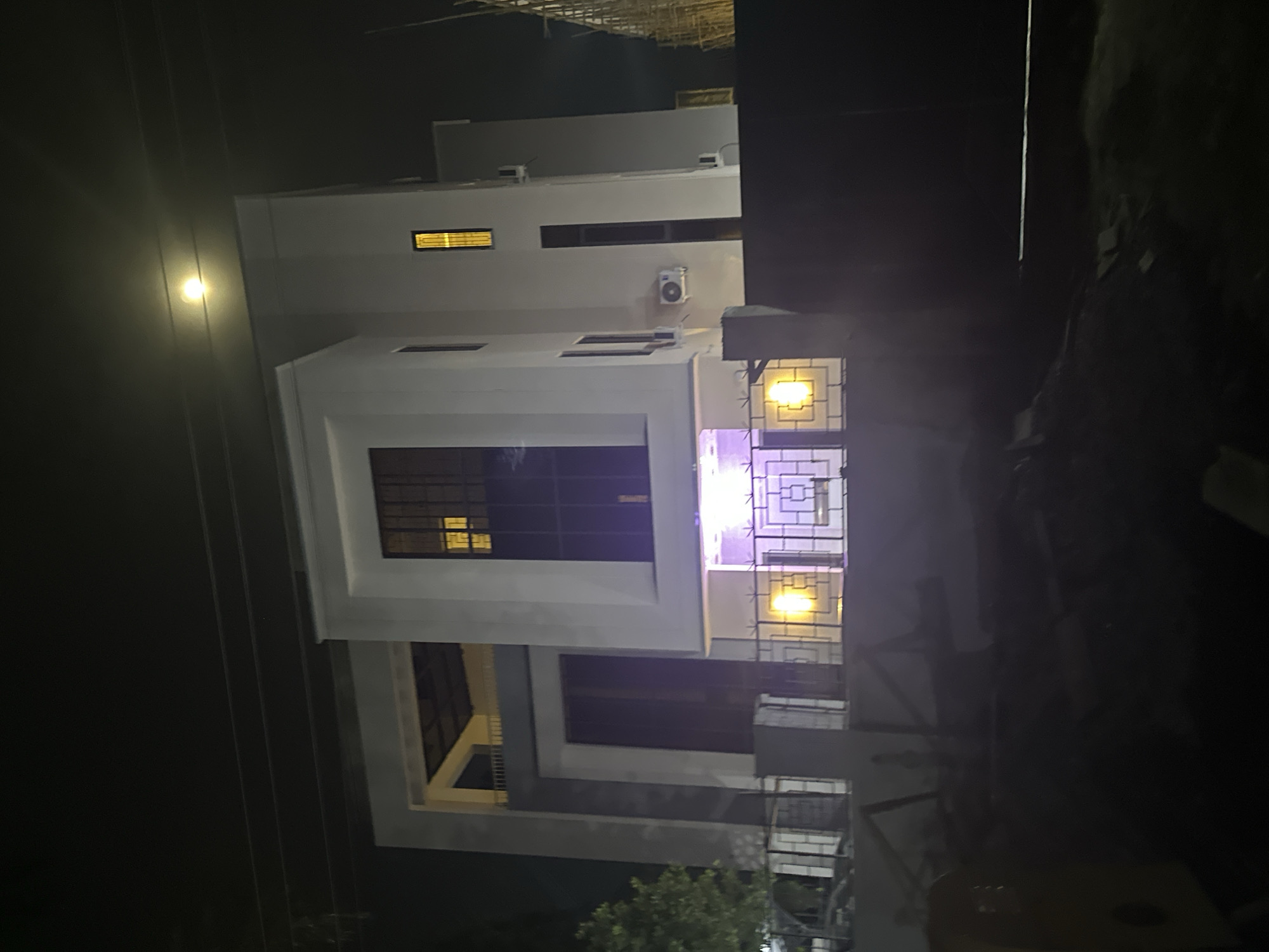 A nighttime exterior shot, illuminated by artificial lights and the moon, captures a contemporary, multi-story white building in Abuja, Nigeria. The building features large, dark-paned windows and several air conditioning units mounted on its exterior walls. A decorative metal fence with a geometric pattern encloses the property, with a dark gate visible to the right. Inside the fence, the ground level is illuminated with warm yellow lights, casting an inviting glow. In the foreground, the scene is cluttered with construction materials and debris, suggesting ongoing development or recent work. The overall impression is one of a modern residence or commercial property under nighttime illumination in a developing urban environment.