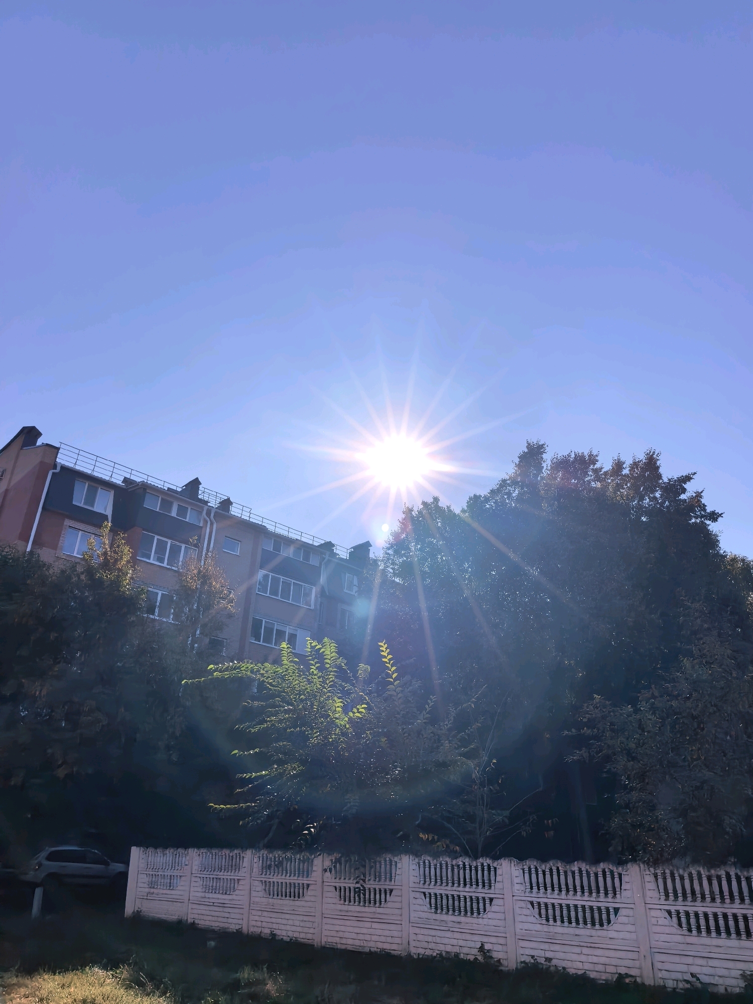 The image shows a residential building in Sterlitamak, Russia, bathed in bright sunlight. The building is multi-story with visible windows and a dark roof. Trees with green foliage surround the building, and a white fence runs along the foreground. A silver car is parked to the left of the frame. The sky is clear and blue, with the sun creating a starburst effect. The scene appears to be an outdoor daytime setting in a residential area.