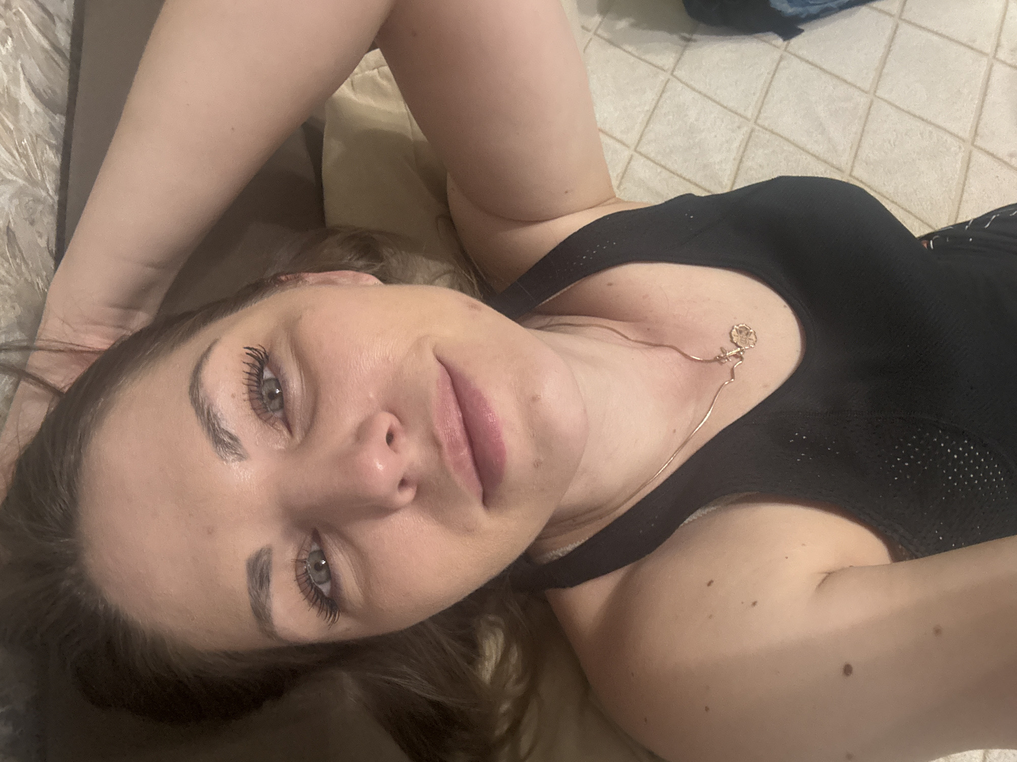 A woman is lying down. She is wearing a black top and a gold necklace. Her eyes are light-colored. The location is not identifiable as Razdolye, Russia. The background includes a patterned floor and bedding.