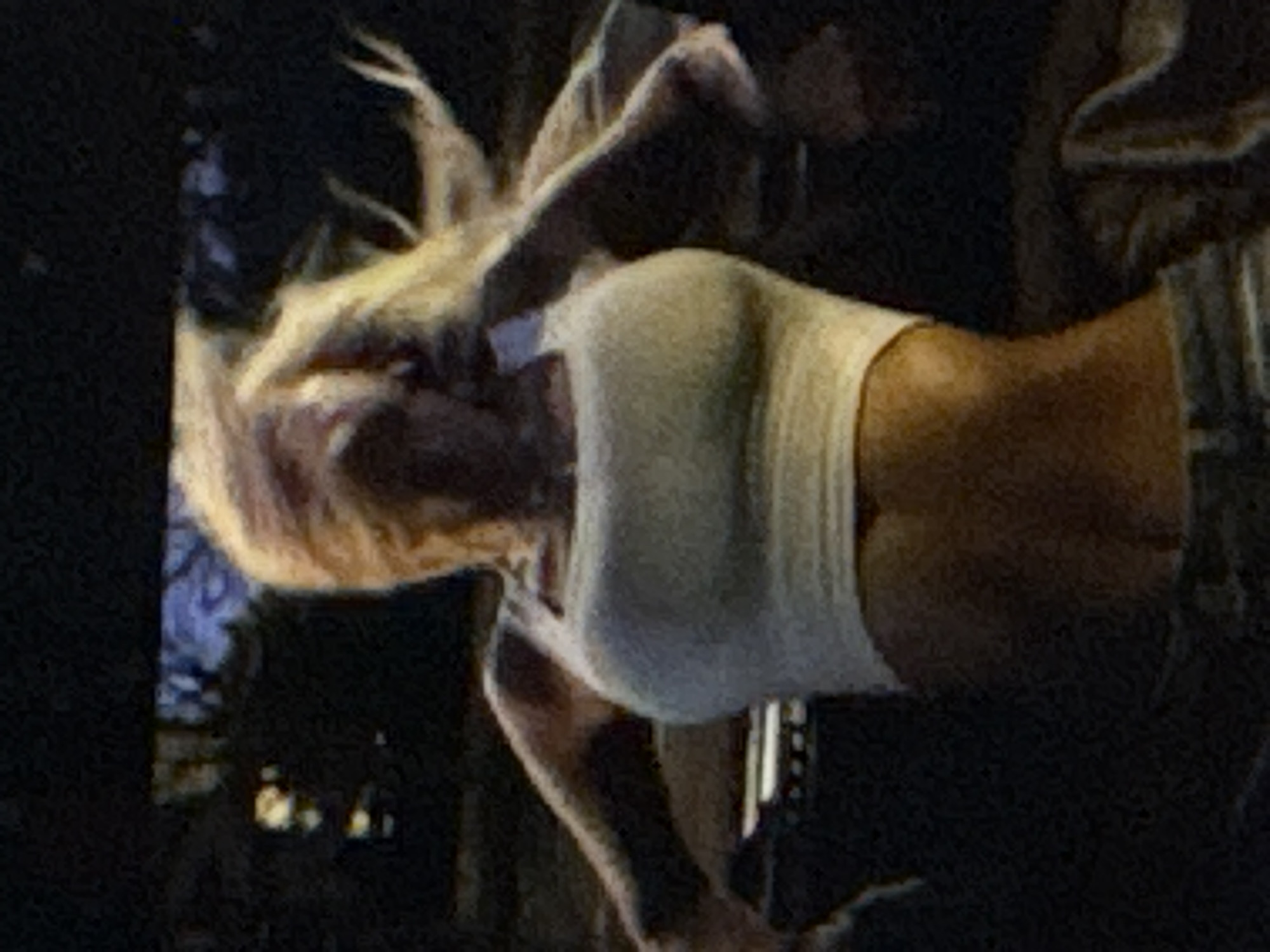The media file shows a close-up, low-resolution image of a person in what appears to be a dark setting, possibly an indoor venue or nighttime. The person has blonde hair that is partially obscuring their face. They are wearing a light-colored, form-fitting top that exposes their midriff. The angle of the shot focuses on the person's torso and arms, with one arm raised and bent at the elbow. The background is dark and out of focus, with some indistinct shapes and lights that could suggest a crowd or stage lighting. The overall impression is of a performance or a candid moment captured during an event. There is no visible text in the image. Given the provided location context of Kuta Selatan, Indonesia, it is plausible this image was taken during a festival, concert, or nightlife event in that area. The lighting suggests it is likely evening or nighttime.
