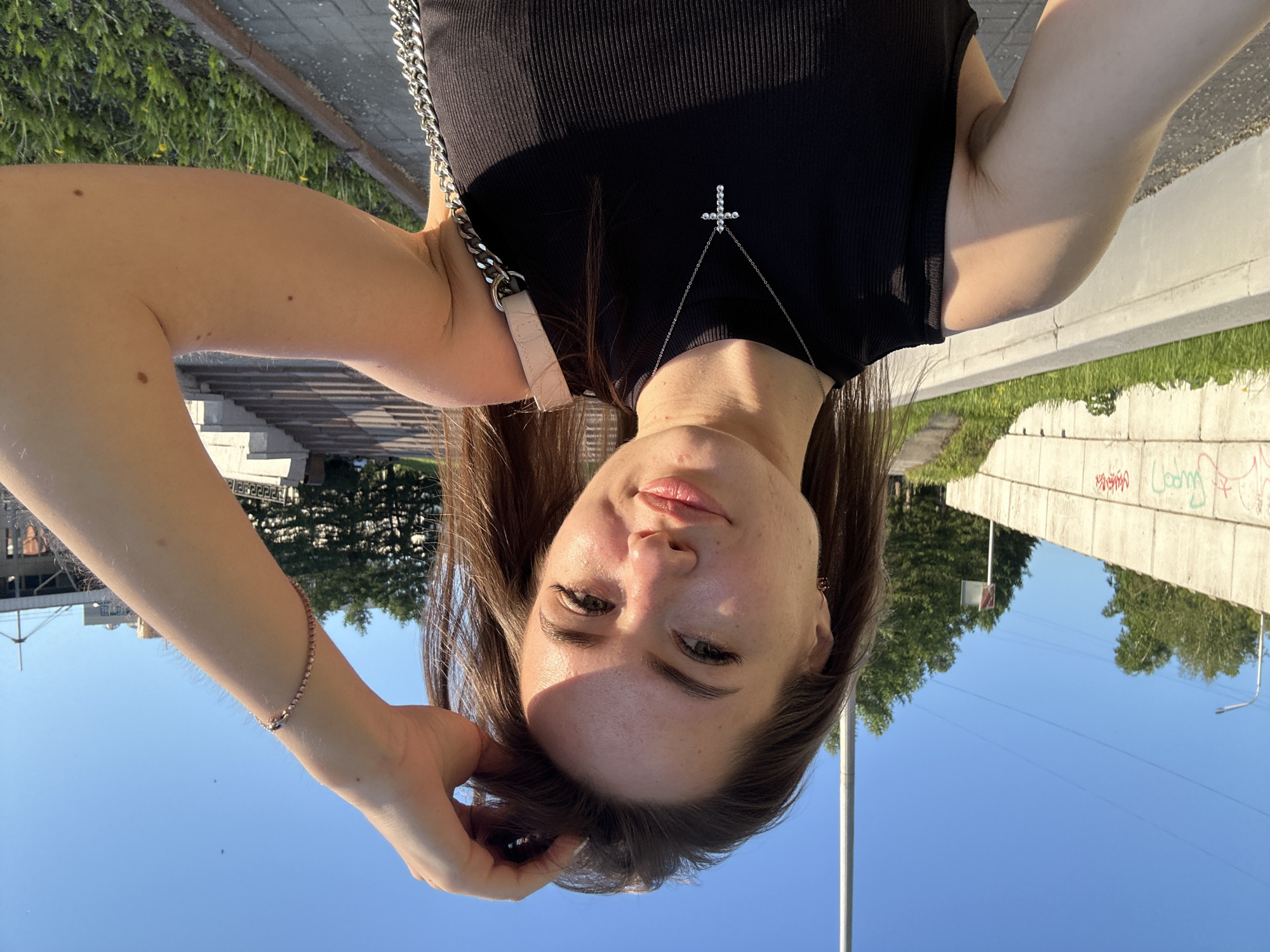 A young woman, appearing to be in her late teens or early twenties with long, dark brown hair, is captured in a close-up outdoor selfie. She is looking directly at the camera with a neutral to slightly smiling expression. The sun casts a shadow across her forehead and nose. She is wearing a black ribbed tank top or sleeveless shirt. A silver chain necklace with a small, sparkly cross pendant rests on her chest. On her left wrist, she wears a delicate, sparkly bracelet. A thick silver chain, likely part of a handbag strap, is visible over her right shoulder.
The setting is an outdoor, urban or suburban environment, likely during the late afternoon or early evening given the clear blue sky and warm, directional sunlight creating shadows. In the background, to her right, there is a concrete wall topped with green grass and trees further behind. Graffiti is visible on this wall, with some letters resembling "coom" or similar illegible scribbles. To her left, there are concrete steps or a similar structure with some greenery. A utility pole or street light is also visible against the clear sky. The image was taken with the camera held above her head, creating an upward-looking perspective.