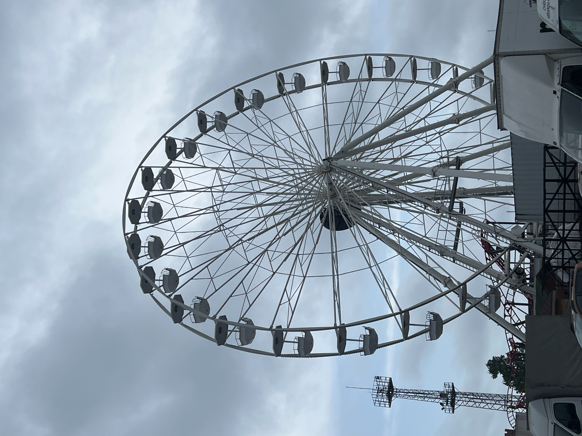 A large Ferris wheel dominates the scene against an overcast, cloudy sky. The Ferris wheel, constructed from white or light-colored metal, features a central hub with numerous radiating spokes supporting its circular rim. Many empty passenger cabins or gondolas are suspended evenly around the rim, suggesting no activity at the moment.
To the bottom right of the image, parts of a large white vehicle or structure, possibly a truck or trailer associated with the fairground, are visible. On its side, some faint, dark text is present, with parts resembling "CREAM" or "KREAM" on a logo, and what might be "ELM" or "EIM" near a window, though the text is not fully legible. Behind this structure and further to the right, a tall, dark metal lattice tower or support structure stands against the sky, with glimpses of dark green foliage visible behind it.
The overall setting suggests a fairground, carnival, or amusement park in Varna, Bulgaria, on a cloudy daytime. No people are visible engaging in activities. The image captures the static structures of the attraction under natural, diffused light.