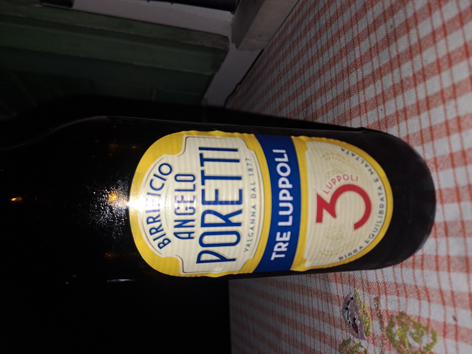 A close-up, slightly angled, eye-level shot showcases a dark glass beer bottle with a prominent label. The label is yellow and blue with white text. At the top of the label, in blue text, are the words "BIRRICICIO ANGELO". Below this, in larger blue text, is "PORETTI". Beneath "PORETTI", in smaller white text, is "VALGANNA DAL 1877". Further down, in a blue band, reads "TRE LUPPOLI". Below this, a yellow band surrounds a red numeral "3". Encircling the numeral "3" is text in a light beige color, reading "BIRRA EQUILIBRATA E MALTATA". The bottle is placed on a table covered with a red and white checkered tablecloth. The background is dark and out of focus, with faint suggestions of a window frame on the right. The scene does not depict any people or specific city locations.