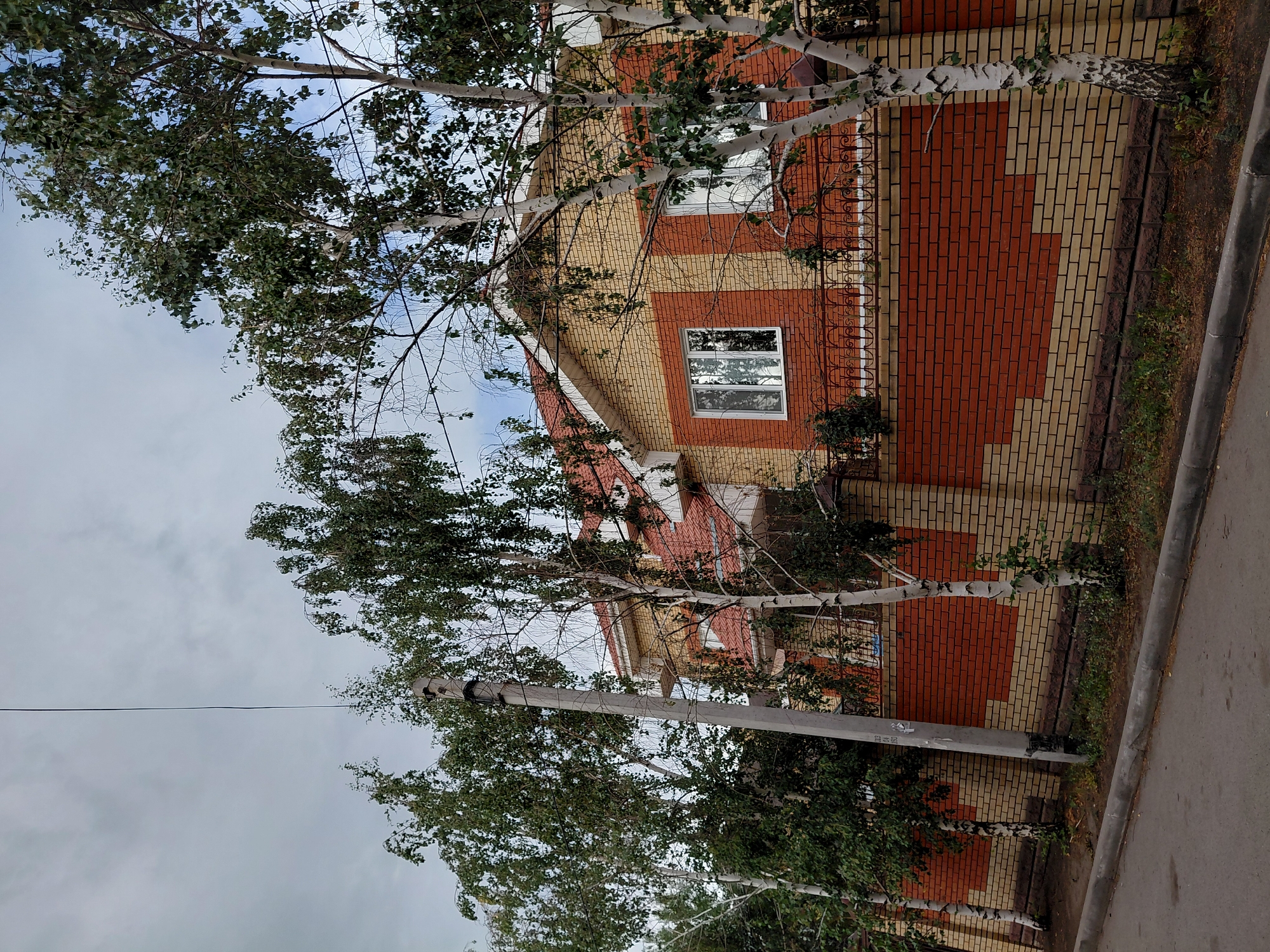A medium-shot, eye-level, outdoor photograph captures a residential property in Qarağandı city administration, Kazakhstan. The house, a two-story structure with a red tiled roof, features yellow and red brick exterior walls. The windows appear to be double-paned with white frames. A decorative metal fence with a curved top runs along the property line, separating it from the street.
Several trees, predominantly birch trees with white bark and green foliage, are scattered throughout the scene, with some branches obscuring parts of the house and fence. A concrete utility pole stands to the left of the frame, with wires extending upwards and across the sky. The ground in front of the fence is covered with grass and some fallen leaves. A paved street with a concrete curb runs along the bottom of the image. The sky is overcast with a mix of grey and lighter clouds. No individuals are present in the image. The scene appears to be captured during daylight hours.
