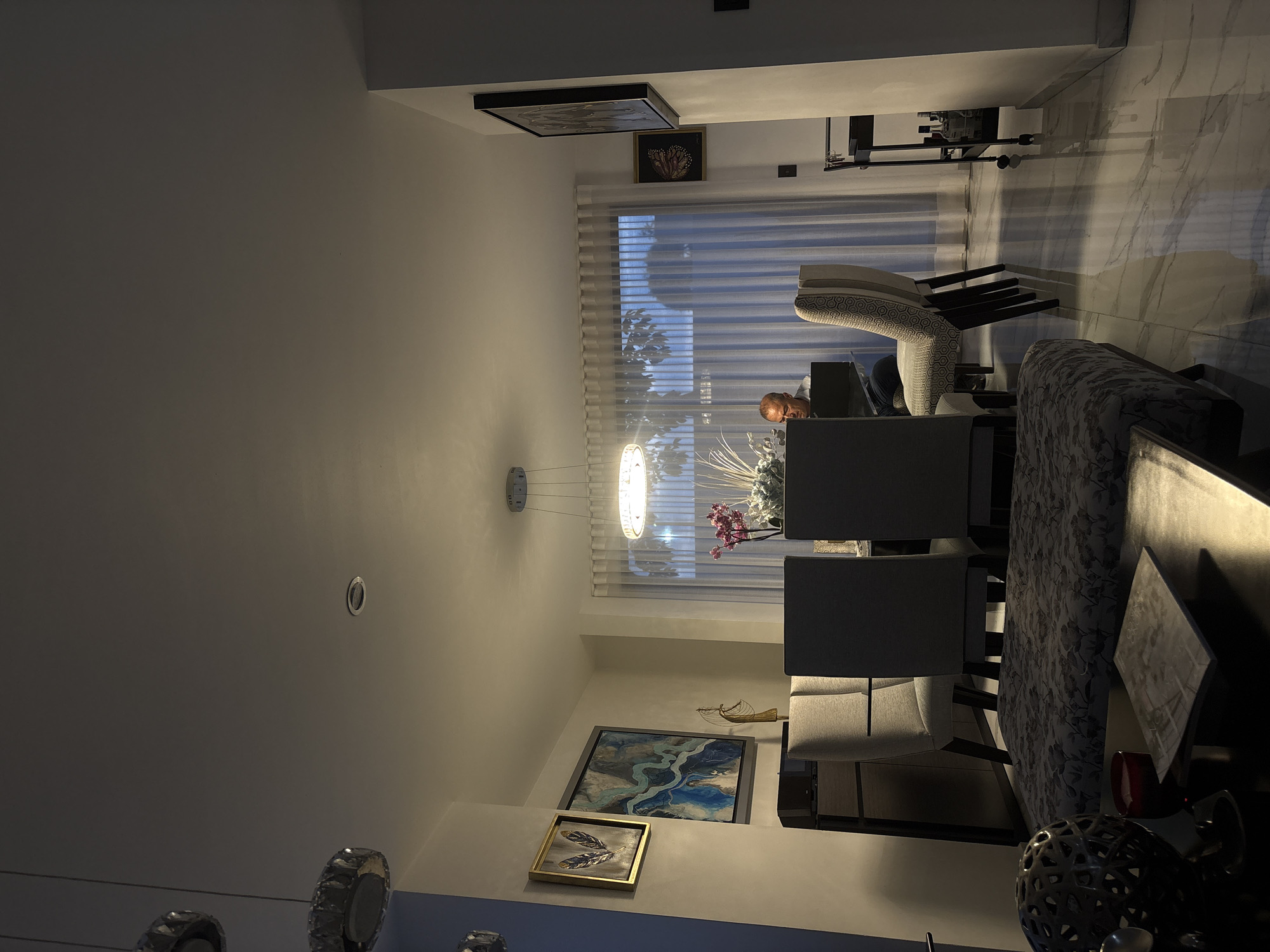 The image, originally captured in a vertical orientation, depicts a modern and elegantly furnished interior space, likely a dining or living area, at what appears to be evening or nighttime. The setting is San Bernardino Tlaxcalancingo, Mexico.
An adult male is partially visible, seated at the head of a dining table, his upper body and face illuminated by light from a laptop screen or similar device. The table itself is partly covered with a dark, floral-patterned cloth. Grey upholstered dining chairs are arranged around the table.
Artificial lighting from a contemporary linear chandelier, suspended above the table, and recessed ceiling spotlights, illuminates the room, creating a warm, serene atmosphere. A large window, covered by sheer, horizontal blinds, shows blurred outdoor foliage, reinforcing the indoor setting during dusk or night.
The walls are adorned with several framed artworks: a prominent abstract painting featuring blue and white river-like patterns, a smaller gold-framed piece depicting golden feathers, and two other framed pictures higher up on the wall. A distinctive marble-patterned wall adds textural interest to the background. Decorative elements include a vase filled with white and pink flowers on the table, and a dark, intricately designed metallic orb on a side surface. The overall scene conveys a sense of quiet relaxation in a stylish home. No visible text is present.