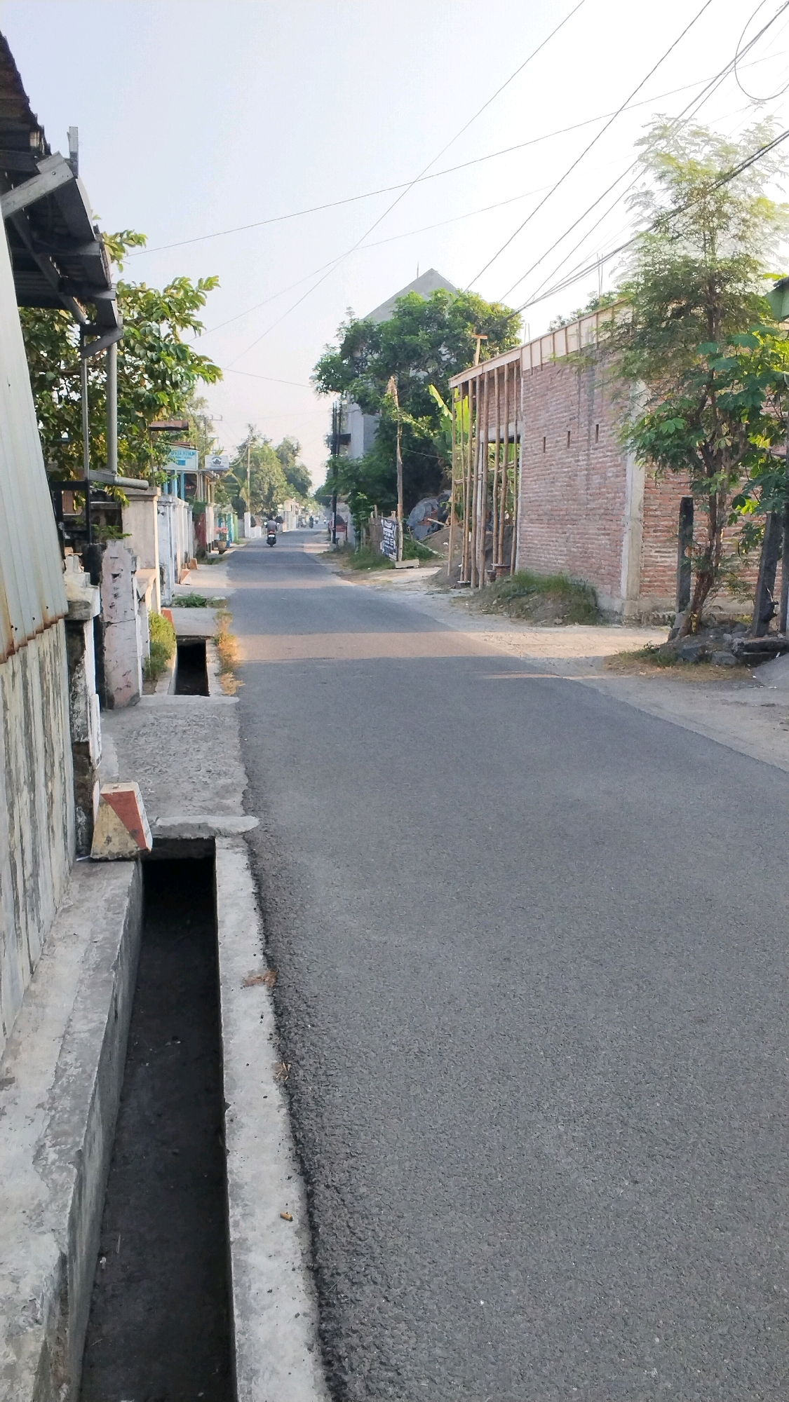 The image shows a street scene in Penompo, Indonesia. A paved road runs diagonally from the foreground to the background, with a concrete drainage ditch on the left. The ditch contains some water and debris. On the right side of the road, a brick wall is under construction, supported by wooden beams. Trees and other vegetation are visible along the sides of the road. In the distance, a motorcycle is being ridden down the street. The sky is clear and the sun is shining, suggesting it is daytime. The overall impression is of a somewhat unfinished or developing urban area.