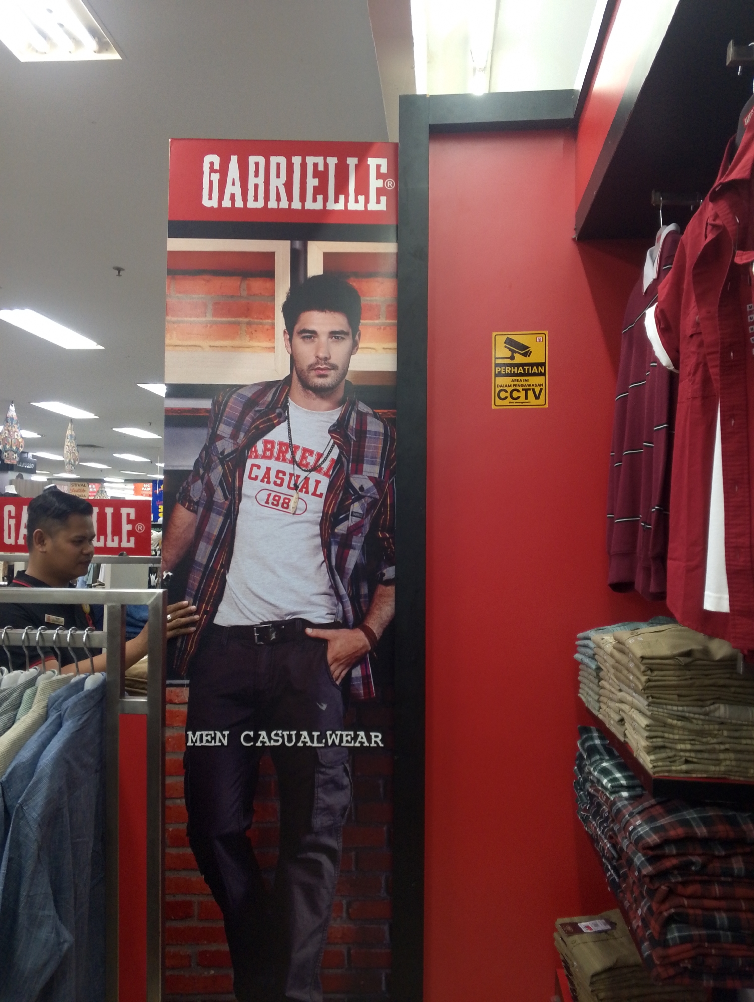 The image depicts the interior of a clothing store, likely in Batam, Indonesia, indicated by the Indonesian text on a CCTV sign. In the foreground, a large advertisement for "GABRIELLE" features a male model wearing a plaid shirt over a t-shirt and dark cargo pants. Text on the advertisement reads "MEN CASUALWEAR." To the left of the advertisement, a store employee, a man with short dark hair, is visible behind a clothing rack. He is wearing a black t-shirt and appears to be looking towards the left. The background shows store shelves stacked with folded clothing, primarily shirts and pants, in various colors and patterns. The store environment is characterized by red walls and bright fluorescent lighting. The CCTV sign is yellow with black text and a camera icon, warning of surveillance.