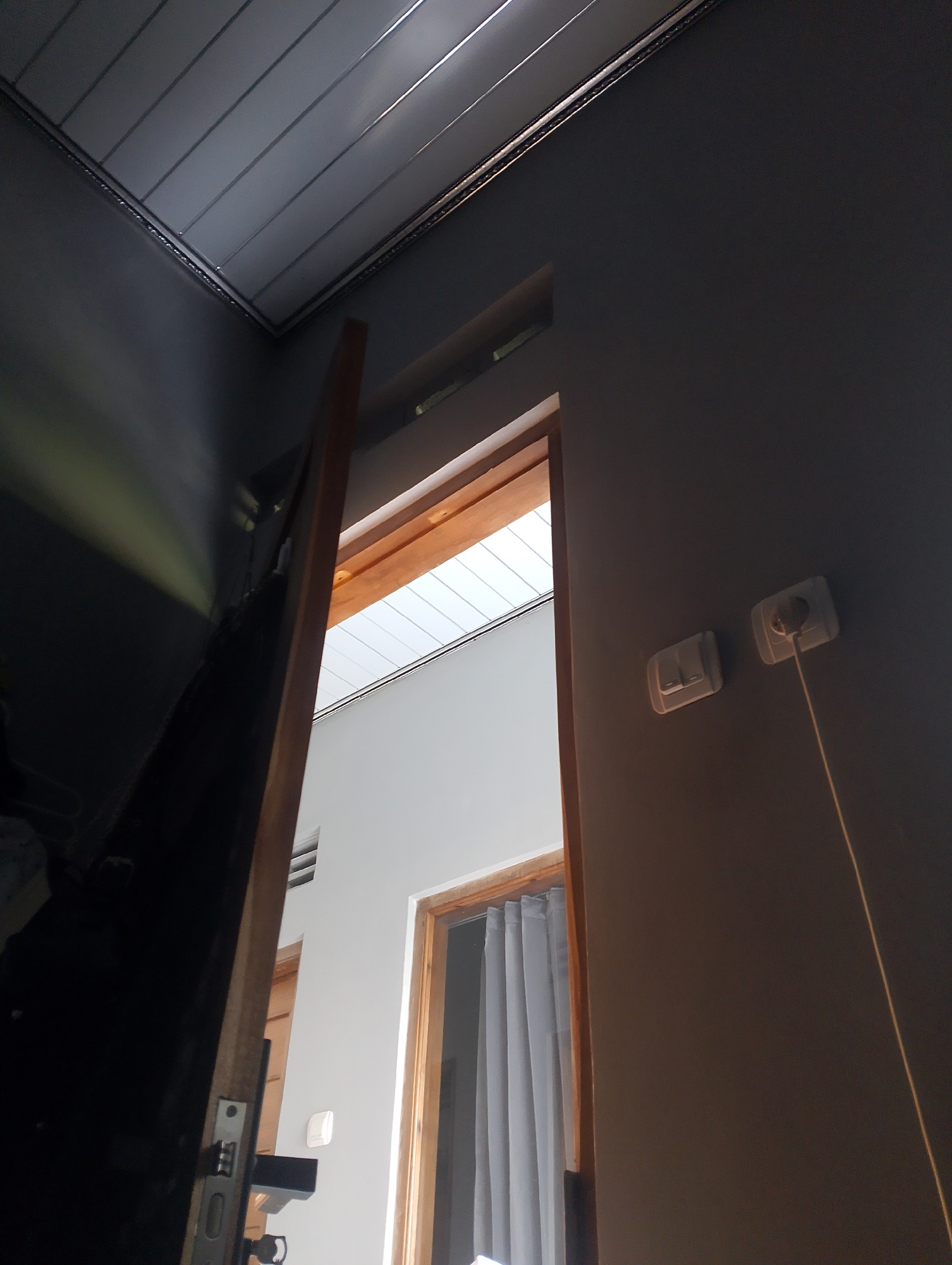 The image shows an interior view from a doorway, looking up and into another room. The foreground is dominated by a dark, reflective surface on the left, possibly a door or furniture, with a wooden door frame angled from the bottom left towards the center. To the right of the door frame, the adjacent wall is painted a dark grey. Two electrical outlets are visible on this wall, with one socket containing a plug connected to a yellow cord.
Beyond the doorway, the room appears to be brightly lit, with a white ceiling composed of horizontal panels. A wooden window frame is visible on the right side of this inner room, with a light grey curtain drawn behind it. The overall environment suggests an indoor space, potentially a residence. The angle of the shot and the lighting suggest the photo was taken during the day. The presence of electrical outlets and switches are standard household features. There are no people or animals visible in the image. The scene does not contain any elements that specifically identify it as being located in Magelang, Indonesia.