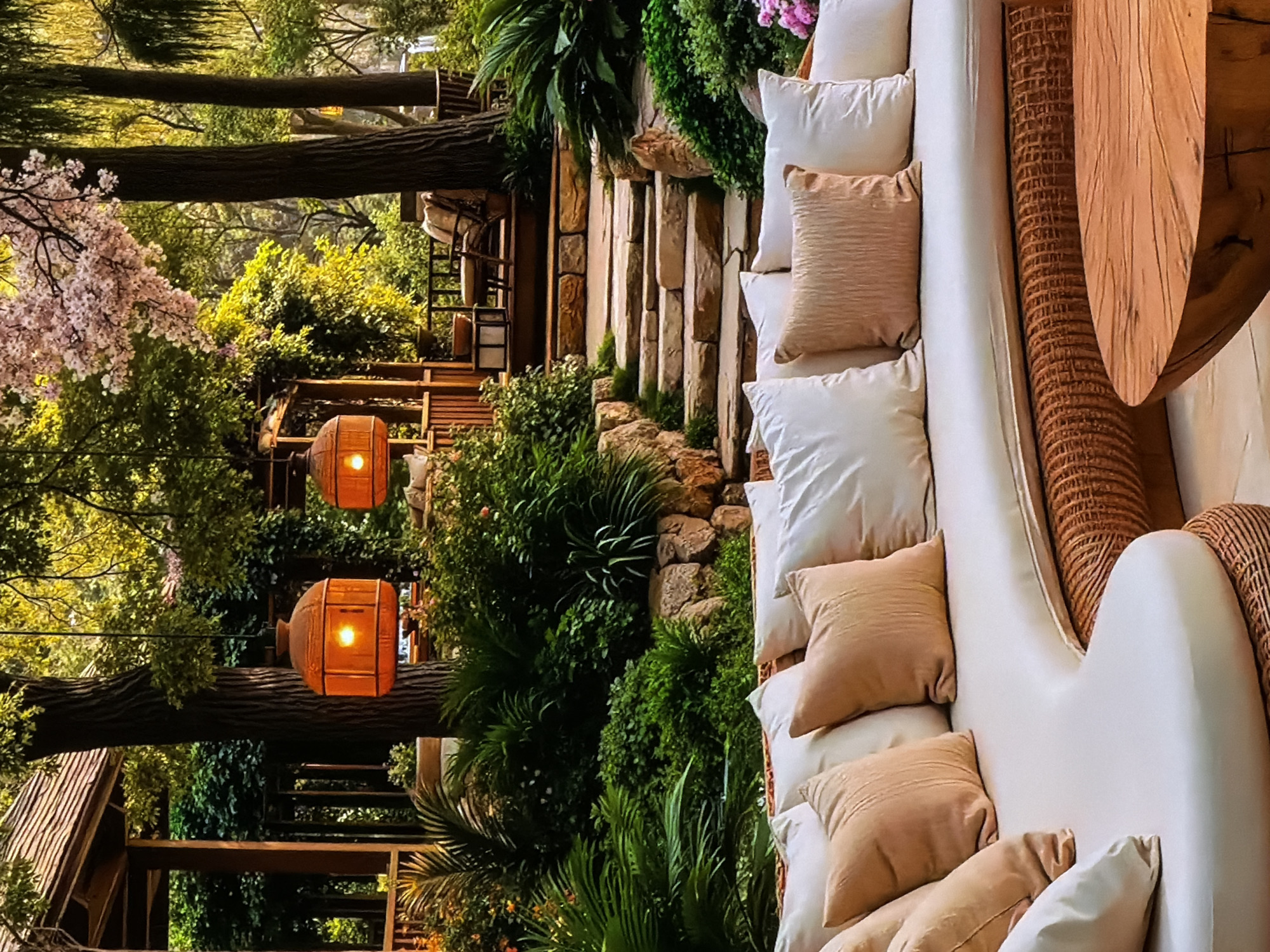 This image captures a lush, inviting outdoor lounge area, likely at a resort or upscale restaurant, in Sofiivska Borshchahivka, Ukraine. The scene is presented with a vertical orientation, but if viewed horizontally, it showcases a serene and green environment bathed in warm light.
In the foreground, a long, light-colored (possibly cream or beige) outdoor sofa is lavishly adorned with numerous pillows. Some pillows are white, while others are a soft, light brown or beige, adding texture and comfort. Adjacent to the sofa, a circular wooden table with a distinctive woven or textured base is visible, suggesting a space for drinks or small meals.
The midground features prominent natural and artificial elements. Two large, cylindrical lanterns, crafted from a warm, orange-toned material, hang from tree branches, their internal lights glowing brightly, indicating late afternoon, dusk, or evening. Behind these, a multi-tiered wooden structure, possibly a deck or pathway, is integrated into the landscape, alongside a rugged stone wall or steps.
The background is dominated by dense, vibrant greenery. Tall, sturdy tree trunks rise vertically, some with spreading branches. A beautiful flowering tree with soft pink blossoms, possibly cherry blossoms, adds a delicate touch of color to the upper left quadrant. Various shrubs, ferns, and lush foliage fill the scene, creating a sense of natural enclosure and tranquility.
No people are visible, emphasizing the peaceful and ready-to-be-enjoyed atmosphere. The overall mood is one of relaxation and luxury, with the warm artificial light contrasting beautifully with the natural light filtering through the trees. No visible text is present in the image.