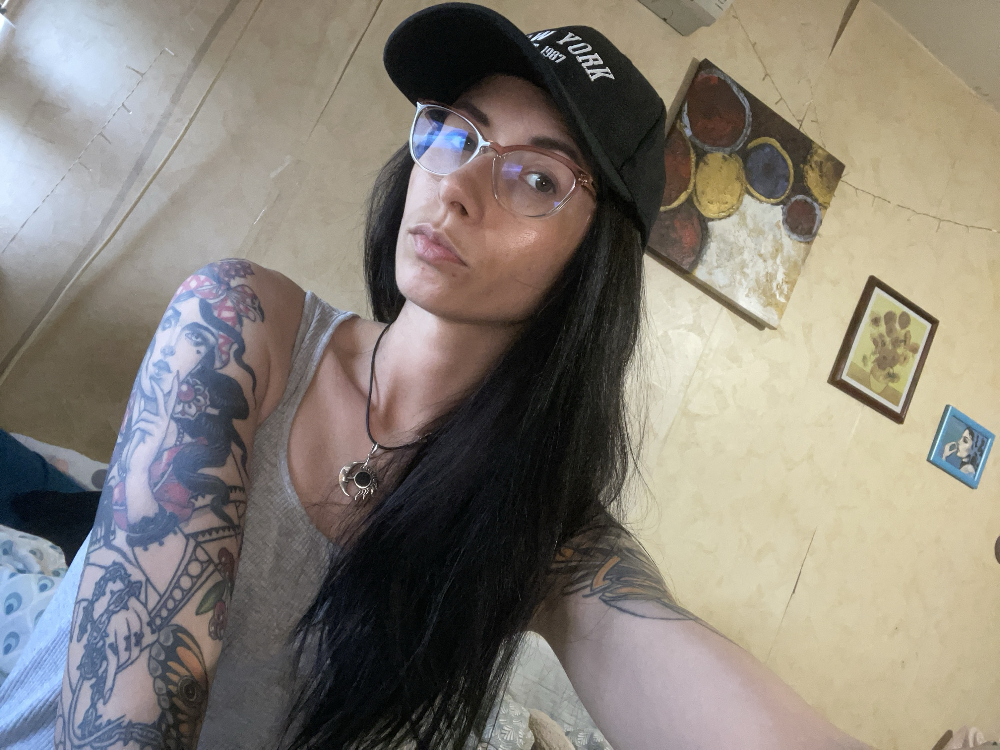 A woman with dark hair and glasses, wearing a black baseball cap with "New York 1987" embroidered on it, is posing for a selfie in an indoor setting. She is wearing a grey tank top and a necklace with a silver pendant. A full-sleeve tattoo is visible on her left arm. The background includes a textured wall with multiple framed pictures and an abstract painting. The city and exact location are unknown.