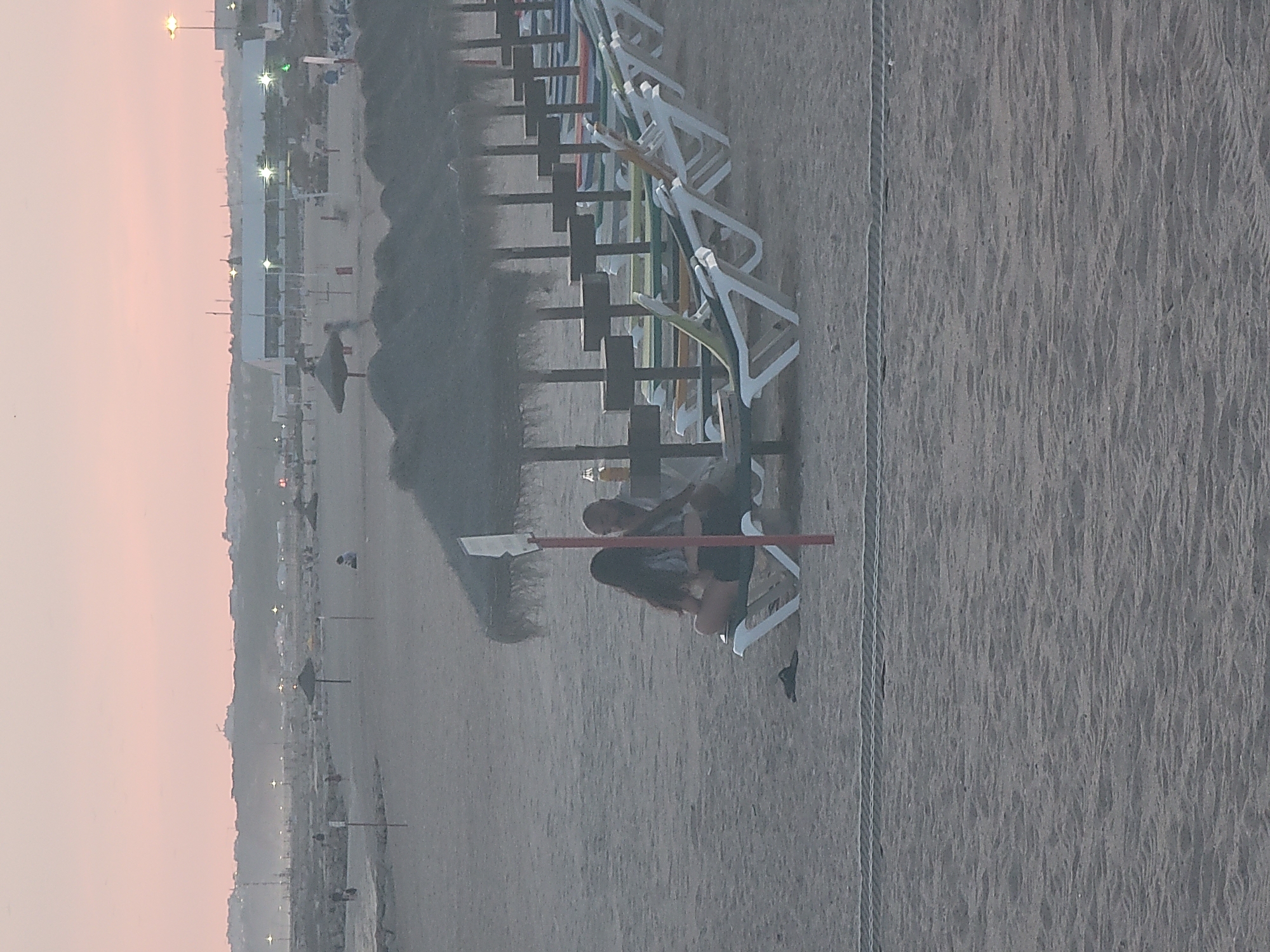 The image depicts a beach scene in Quarteira, Portugal, during what appears to be dusk or early morning, with a soft, pinkish sky. In the foreground, the sandy beach stretches out, with a rope barrier running horizontally across the bottom of the frame. Several rows of empty, white plastic sun loungers are visible, partially covered by thatched umbrellas, suggesting a resort or public beach area.
Further back, two individuals are seated on a sun lounger under one of the thatched umbrellas. A woman with long, dark hair is sitting with her legs crossed, facing towards a man who is seated next to her. A bottle with a yellow liquid, possibly a drink, is placed on a small table or surface between them.
In the mid-ground, more sun loungers and umbrellas are scattered across the beach. In the distance, a hazy horizon shows a cityscape with buildings and lights. Several lampposts are also visible, illuminating the area. Some indistinct figures can be seen walking or sitting on the beach in the further distance. The overall impression is a quiet, relaxed atmosphere on a beach at the end of the day.