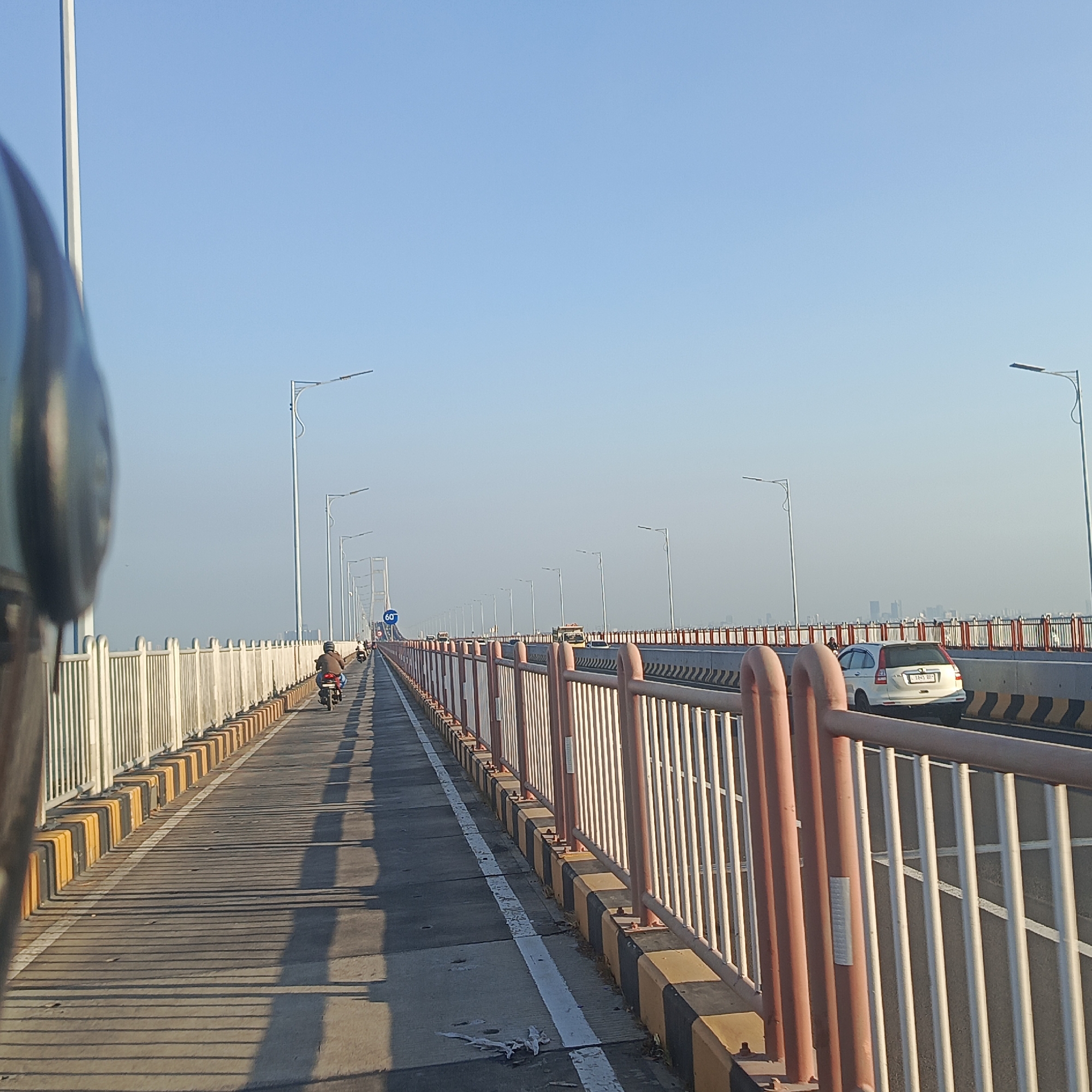 This is a street-level, eye-level shot captured from a vehicle, looking forward along a bridge with a clear blue sky overhead. The bridge has a distinct guardrail consisting of white metal poles and reddish-orange metal poles, interspersed with yellow and black striped barriers along the edge. The road surface is paved, with white lane markings visible.
In the foreground, on the left, a portion of the vehicle's side mirror and its frame is visible, slightly out of focus. A single motorcyclist, wearing a helmet and a brown jacket, is riding away from the camera on the left side of the road. Further down the bridge, other vehicles, including a white SUV, are visible, and a speed limit sign indicating "60" is present. The bridge appears to be part of a larger infrastructure, with multiple streetlights lining its sides, casting long shadows on the road due to the low angle of the sun. In the far distance, a city skyline is faintly visible through the haze. The overall impression is of a journey across a modern bridge on a clear day.