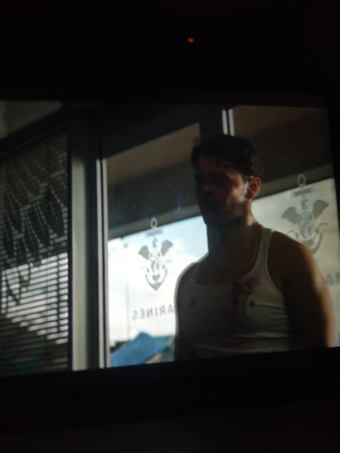 A man in a white tank top stands in front of a glass door. The door has a logo with an anchor, wings, and an eagle. The word "MARINES" is written in reverse. The man has a slight beard and appears to have a mark or stain on his chest, possibly blood. A window with blinds is visible to the left of the door. The overall image is dimly lit, as though taken from a TV screen or monitor.