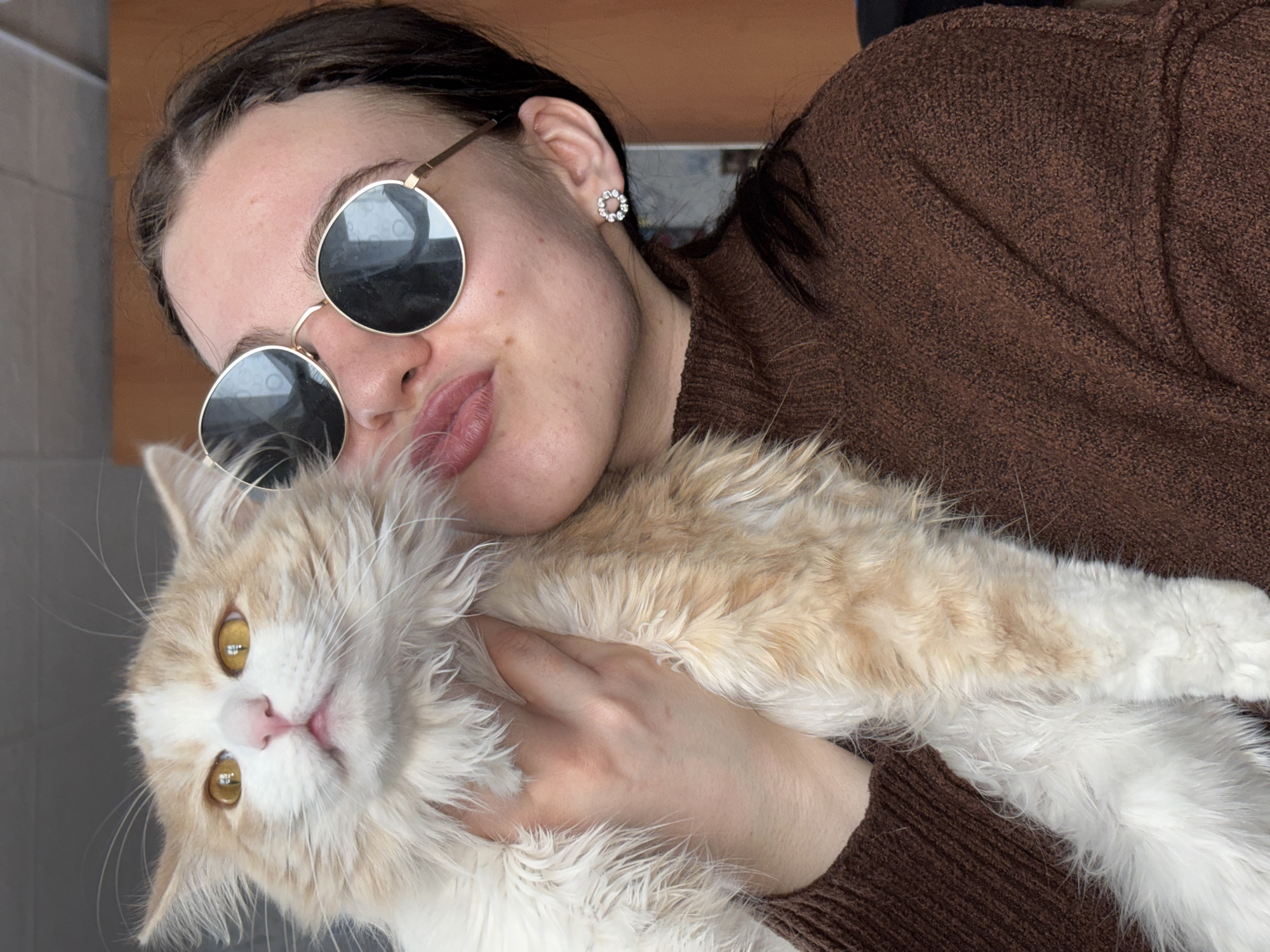 A woman with dark hair pulled back and wearing round sunglasses is holding a light-colored, long-haired cat. The woman is wearing a brown sweater and a sparkly earring is visible in her left ear. The cat has amber eyes and a pink nose. The location is inferred to be in Zasechnyi, Russia. The woman is holding the cat close, with her hand on its chest. The background is out of focus, but appears to be an indoor setting.