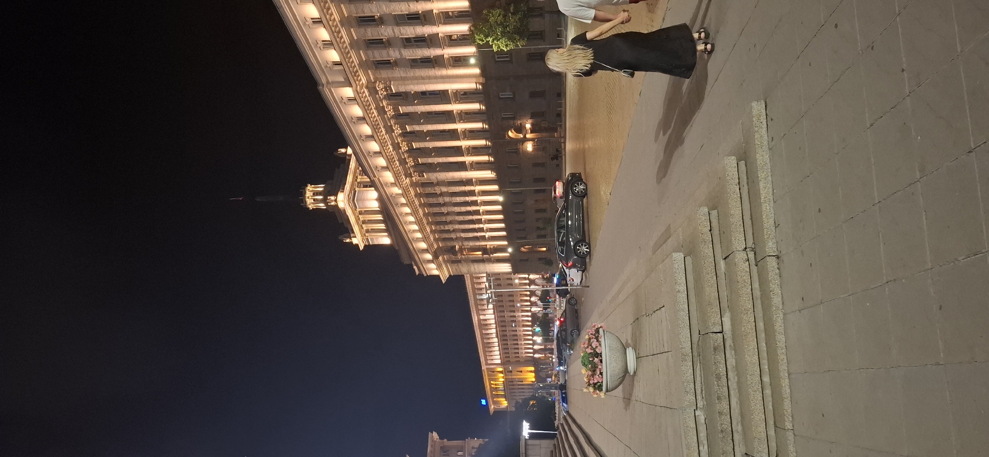 This is a vertical, low-angle nighttime photograph of a grand, classically styled building illuminated by warm lights. The building features multiple stories with rows of windows, cornices, and decorative columns. In the foreground, to the right, is a paved walkway made of large stone tiles, with a series of concrete planters also made of stone tiles lining the edge.
On the walkway, a person with long, blonde hair wearing a long black dress and sandals is walking away from the camera, holding hands with another person who is mostly out of frame. Further down the walkway, a dark-colored car is parked on the street, and other vehicles, including what appears to be a police car, are visible. A large, ornate planter filled with colorful flowers is situated on the walkway. The sky is dark, indicating it is nighttime. The overall scene suggests an urban setting, possibly a public square or a street in front of an important building, with a sense of evening leisure or activity. The architecture of the building and the style of the paving stones provide clues to a European location. The lighting creates a dramatic effect, highlighting the building's features against the dark sky.
