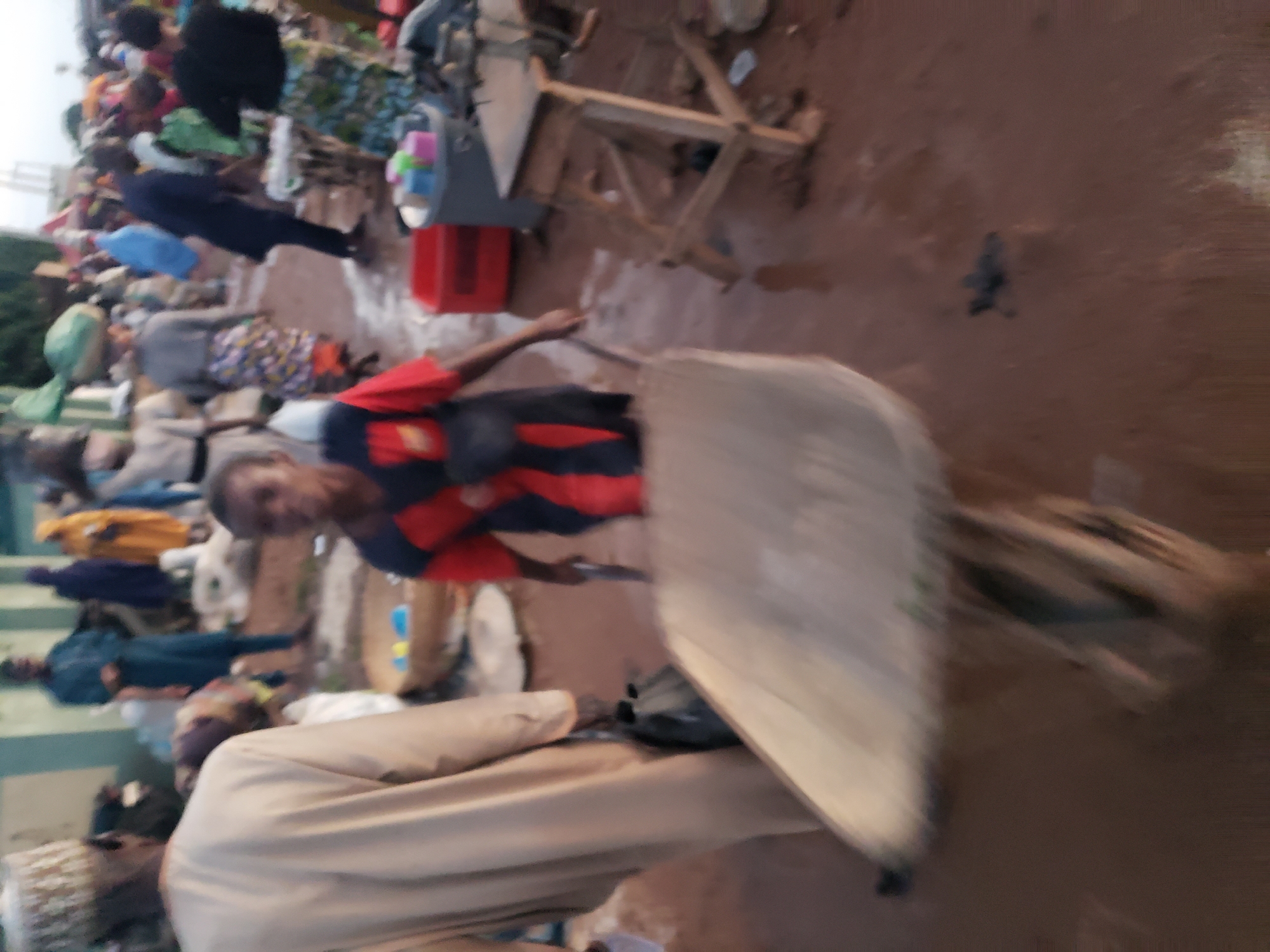 The image depicts a street scene in Kakuri Gwari, Nigeria. In the foreground, a person wearing a tan robe and a cap is partially visible from the back, carrying a black bag. To the right and slightly behind, a child in a red and black shirt is pushing a wheelbarrow. The ground is wet and muddy, suggesting recent rain or a water source. In the background, other individuals are present, some carrying loads on their heads and others engaged in activities within what appears to be a market setting. Various objects, including baskets, crates, and containers, are scattered around. The overall atmosphere is that of a bustling local marketplace.